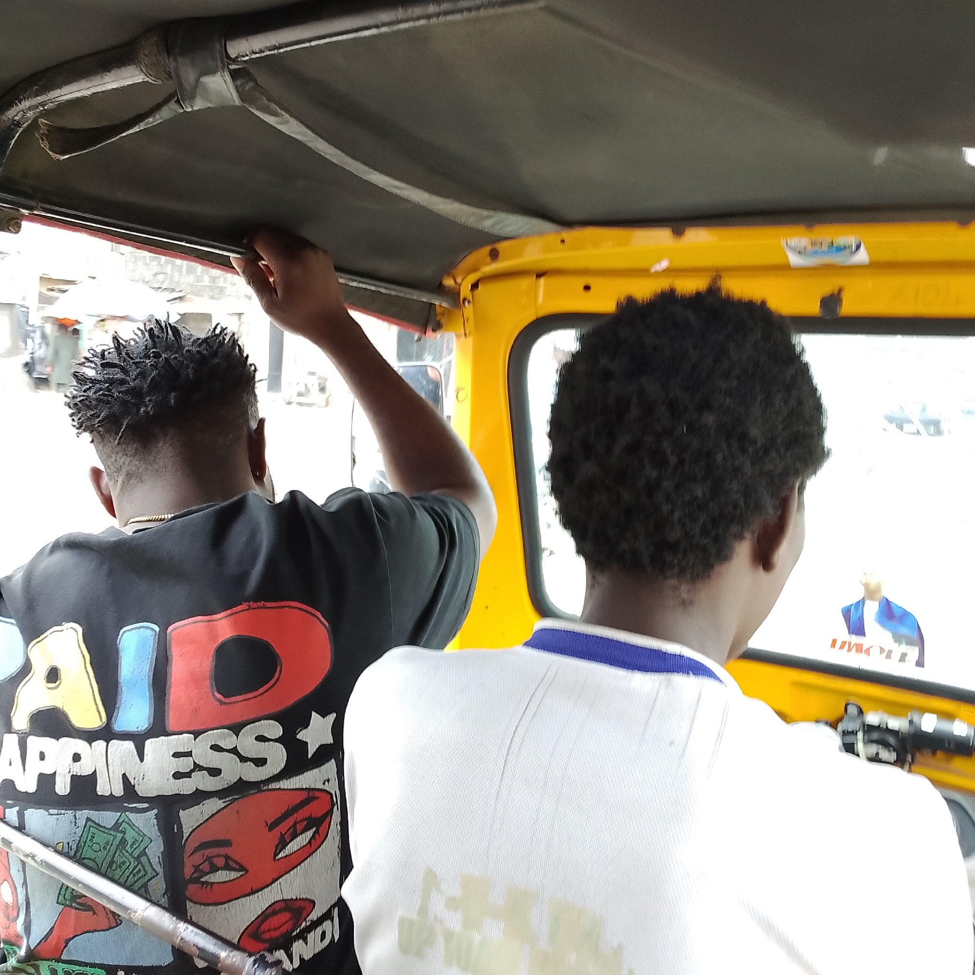 Two individuals, viewed from behind, are seated inside a yellow, open-air vehicle, possibly a motorized rickshaw or tuk-tuk, traveling through Ado, Nigeria. The person on the left wears a black t-shirt with a colorful graphic design that includes the words "PAID HAPPINESS" and imagery of money and lips. This individual has dark, dreadlocked hair and is reaching up to touch the vehicle's roof. The person on the right has dark, curly hair and wears a white polo shirt with a blue collar. The interior of the vehicle shows a black headliner with visible metal framing, and the yellow exterior is visible around a window. Outside the vehicle, a blurred street scene is visible, suggesting an urban environment. The lighting indicates it is daytime.