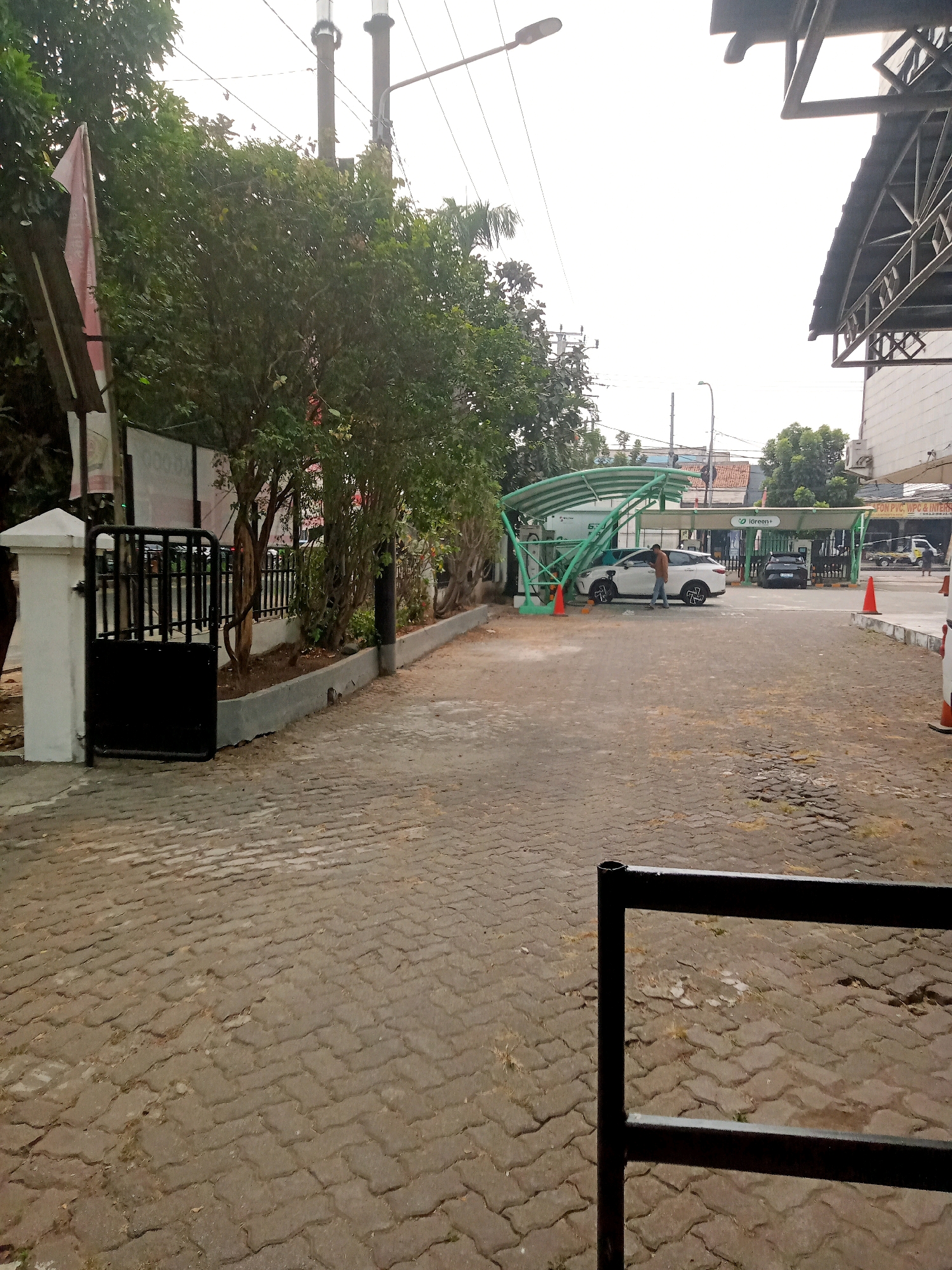 The image shows an outdoor view of a charging station for electric vehicles in Bekasi, Indonesia. In the foreground, a dark metal gate or barrier is visible, partially obscuring a paved walkway made of interlocking concrete blocks. To the left, a white pillar supports a black metal gate, and beyond that, lush green trees line the property. Further back, a white electric car is parked under a green and white canopy structure, connected to charging equipment. A person in a brown shirt stands near the white car. Another black electric car is parked further down under a similar canopy. Orange traffic cones are placed around the charging area. The environment suggests a commercial or public service area with overhead electrical wires and streetlights visible in the background, along with buildings and more trees. The sky is overcast.