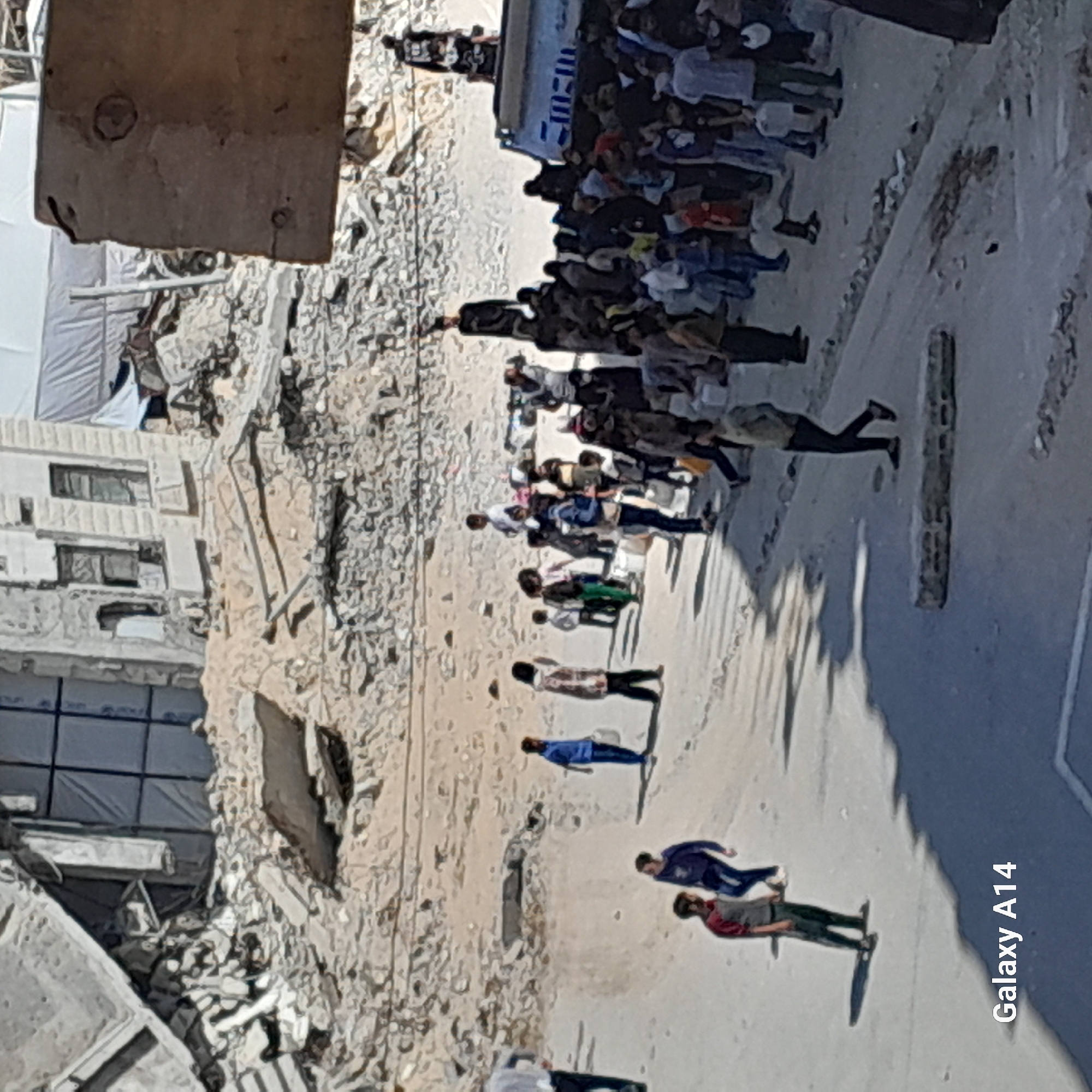 A medium shot captures a street scene in what appears to be a war-torn urban area in an unknown city. The image shows significant destruction on the left side of the frame, with crumbled buildings and debris scattered. On the right side of the frame, a long line of people extends towards a white van parked further away. Some individuals are wearing masks. The ground is visible and appears dusty or sandy. The overall impression is one of devastation and displacement.