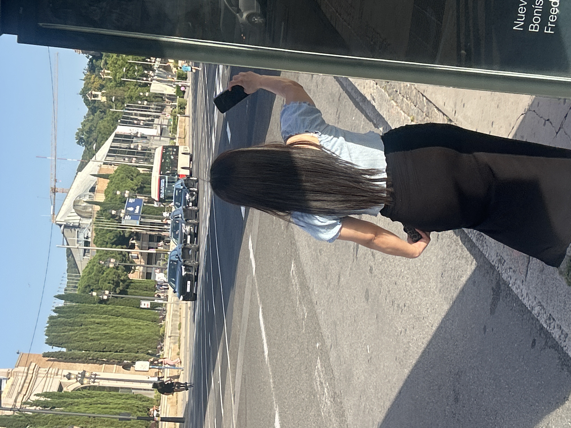 A woman with long dark hair, wearing a light blue off-the-shoulder top and a black skirt, stands at a bus stop on a sunny day in Barcelona, Spain. She is holding up a dark-colored smartphone in her right hand, seemingly taking a photo or video. In the background, traffic moves along a wide street with multiple lanes. Several cars, including black sedans and a white bus, are visible. A cyclist is seen on the left side of the street. The environment includes architectural elements with arched facades, tall cypress trees, streetlights, and a clear blue sky. The bus stop appears to have a glass enclosure with some text on it, including the words "Nuev", "Bonis", and "Freed". The scene is framed by the edge of the bus stop structure on the right.