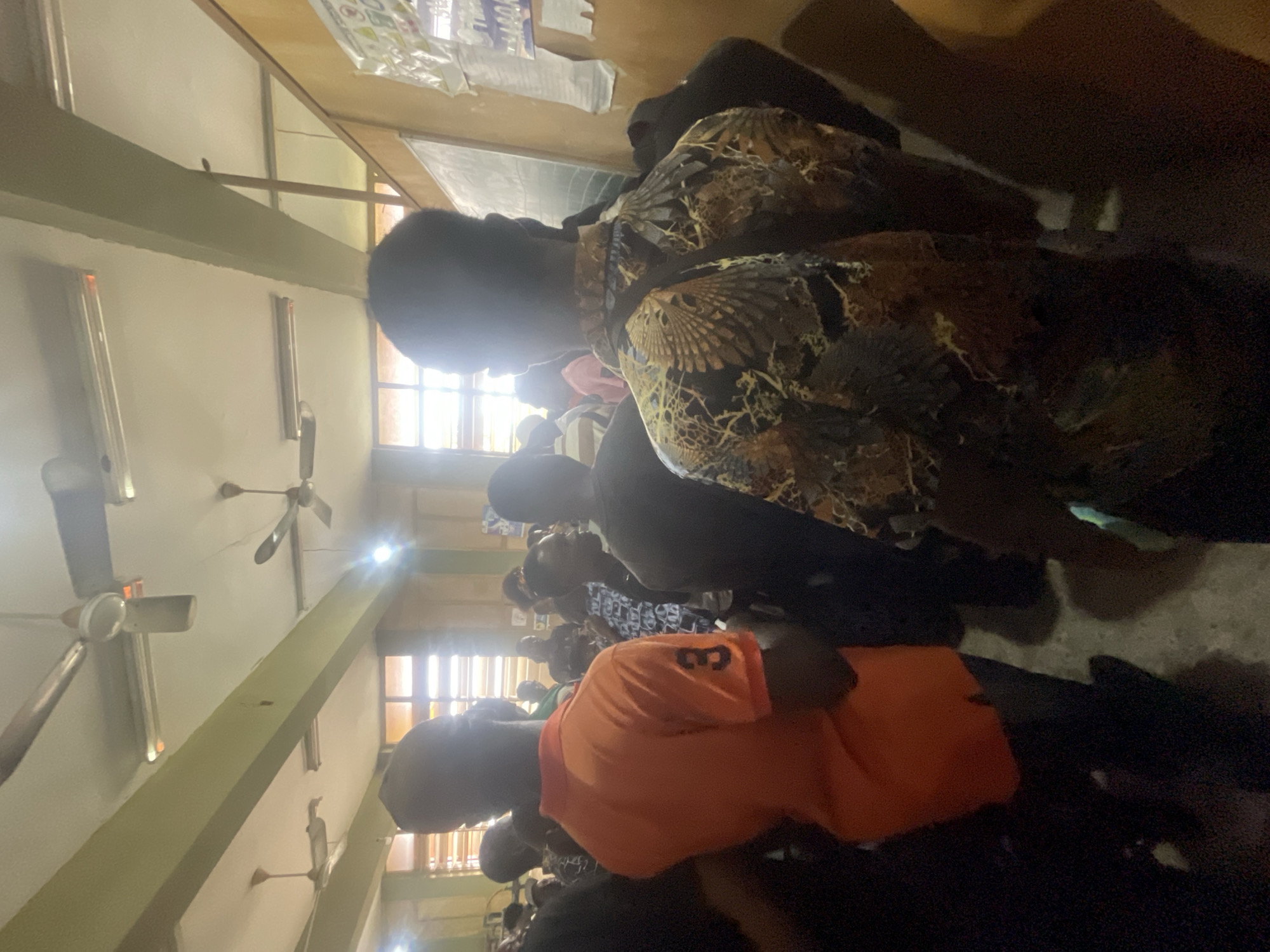 The image depicts an indoor gathering, likely a classroom or meeting hall, with fluorescent lighting and ceiling fans visible. Several individuals are present, mostly viewed from behind. In the foreground, a person wears an orange polo shirt with the number "3" on the sleeve. To their right, another individual sports a patterned shirt with what appears to be avian motifs. The background shows more people and a chalkboard on the wall, suggesting an educational or communal setting. The overall atmosphere appears to be one of social interaction or an event. The location is identified as Ihiagwa, Nigeria.