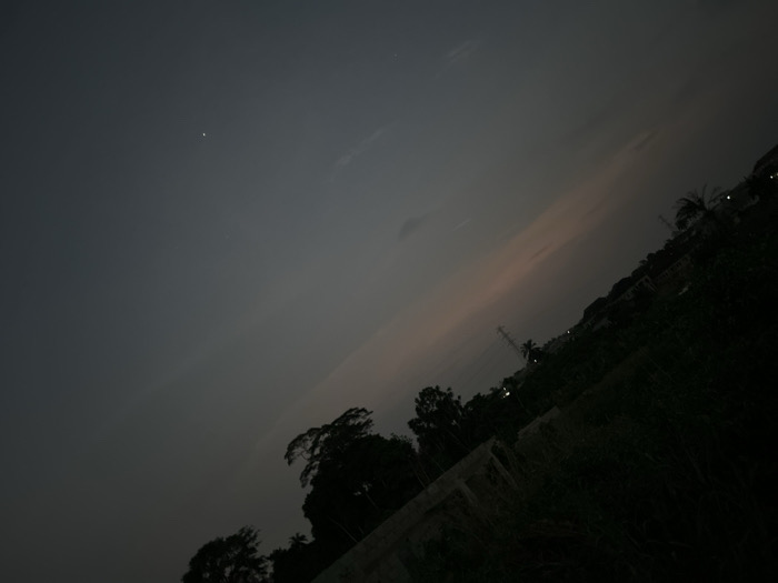 The image shows a nighttime landscape with a dark, overcast sky. A single bright star or planet is visible. Silhouetted trees and vegetation dominate the lower portion of the frame. Faint hints of a building structure are present. In the distance, faint lights suggest a residential area. The sky shows a gradient of light towards the horizon, hinting at a sunset or twilight.