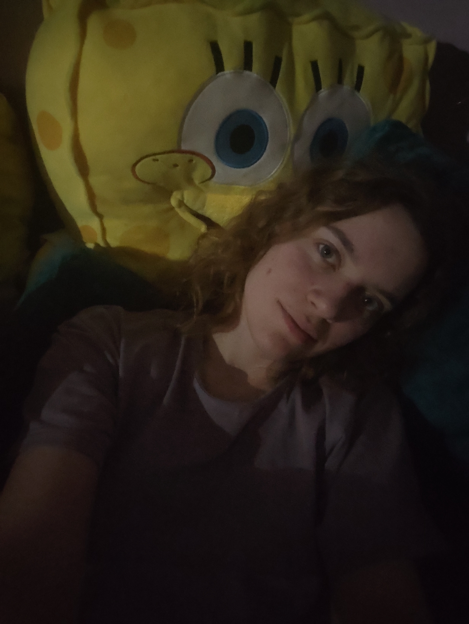 A person with curly, reddish-brown hair is in the foreground. The person is wearing a dark-colored shirt. Behind and slightly above the person is a large, plush toy of Spongebob Squarepants. The toy is yellow with large, cartoonish eyes and a small, round nose. The environment is dark, with a dark blue pillow or cushion behind the person and Spongebob toy. The location is Unknown, Unknown.