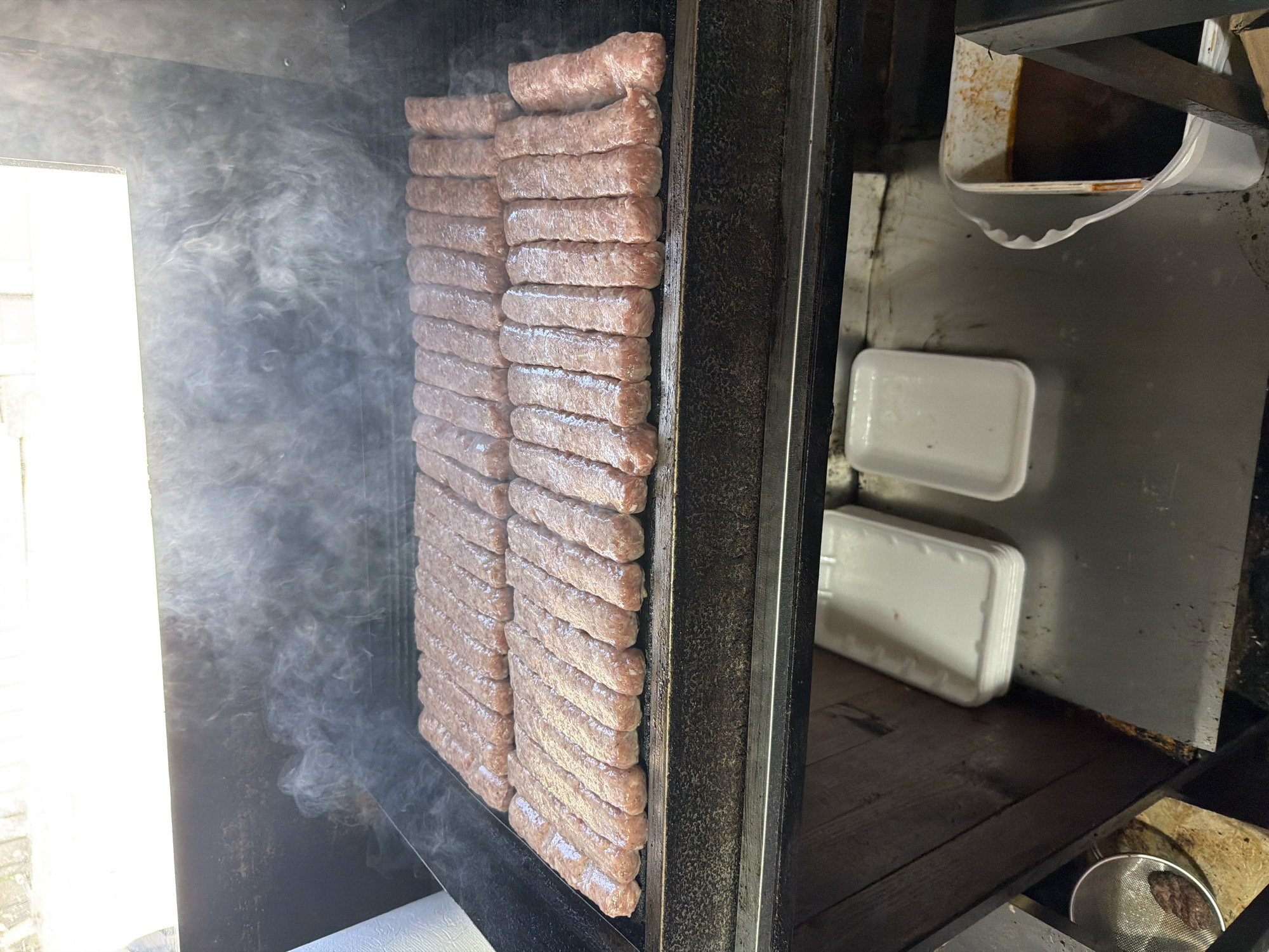 The image displays a close-up view inside a dark metal cooking apparatus, likely a grill or smoker, with numerous raw meat products being prepared. On the left side of the frame, thick white smoke billows outward from the cooking chamber towards a brightly lit, open area, indicating active cooking.
The central subjects are multiple rows of elongated, cylindrical, light pinkish-brown meat items, resembling cevapi or similar Balkan-style sausages. They are stacked vertically in tightly packed columns against a dark metal rack or grate. The meat appears moist and uncooked or only minimally cooked.
The right side and bottom of the frame reveal more of the cooking environment. The surfaces are dark, possibly metal, exhibiting signs of heavy use, including some rust and grime. Several white, rectangular plastic containers or trays are visible; some are stacked neatly, while one appears positioned below the meat to collect drippings or grease. A metal strainer containing some dark residue rests on a lower surface.
The activity depicted is the large-scale preparation and cooking of meat, likely in an outdoor or semi-outdoor setting. The bright light on the left suggests it is daytime. The style of meat product strongly hints at a traditional Balkan culinary context, consistent with the given location of Belashtitsa, Bulgaria. No text is visible in the image.