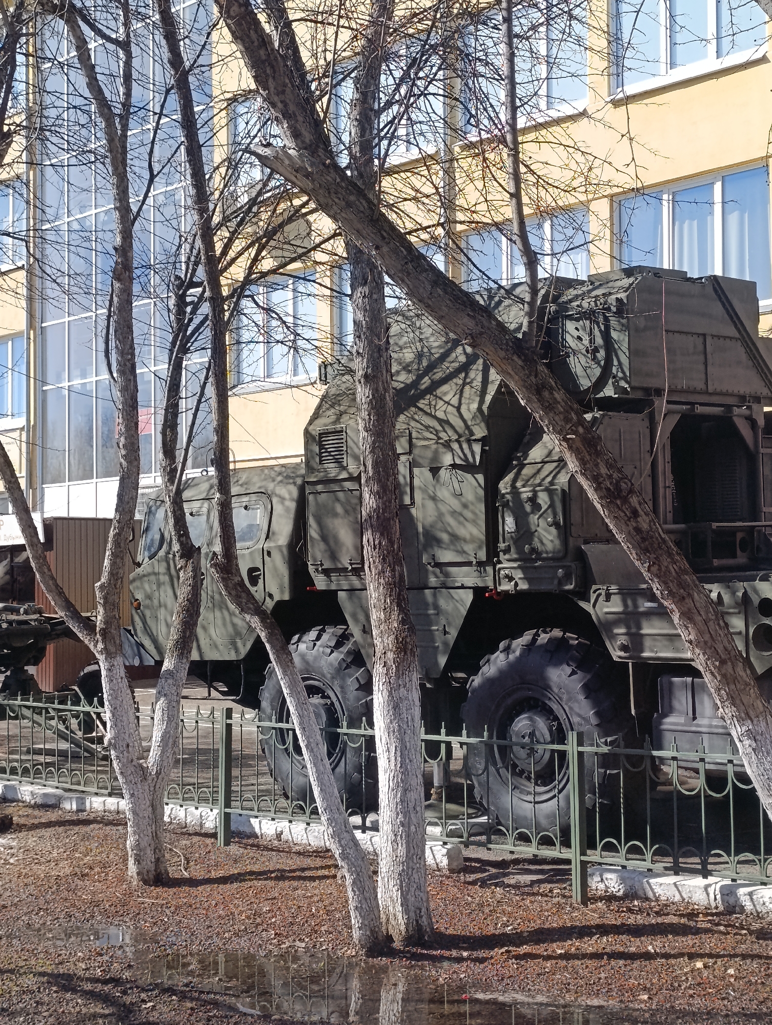 The image shows a large, olive green military vehicle parked behind a dark green metal fence. Several bare trees with white-painted lower trunks stand in the foreground, partially obscuring the view of the vehicle. The vehicle has large wheels and what appears to be a complex system of equipment mounted on the back. In the background, there is a yellow building with many windows. The ground is covered in brown leaves and wet patches, suggesting recent rain or melting snow. The location is Krasnoyarsk, Russia, as indicated by the image's original title or source. The scene is a static display of military equipment.