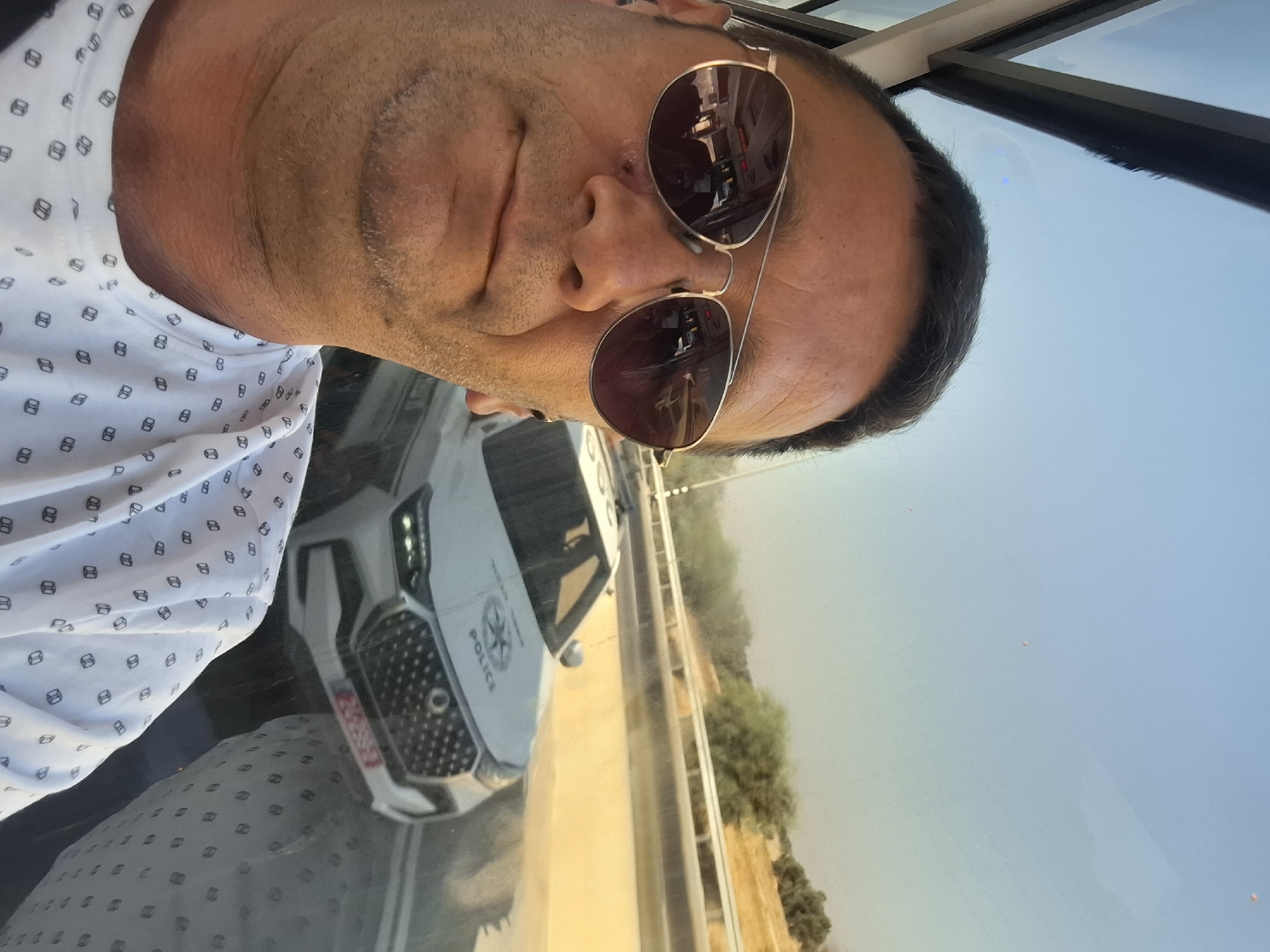 A man wearing sunglasses and a white t-shirt with a black repeating pattern takes a selfie inside a vehicle. A police car with the word "POLICE" on it is visible outside the vehicle through the window. The police car has a license plate with red characters. The scene appears to be on a road in a rural area with vegetation and a blue sky visible in the background. The image was taken in the city of Bnei Shimon Regional Council, Israel.