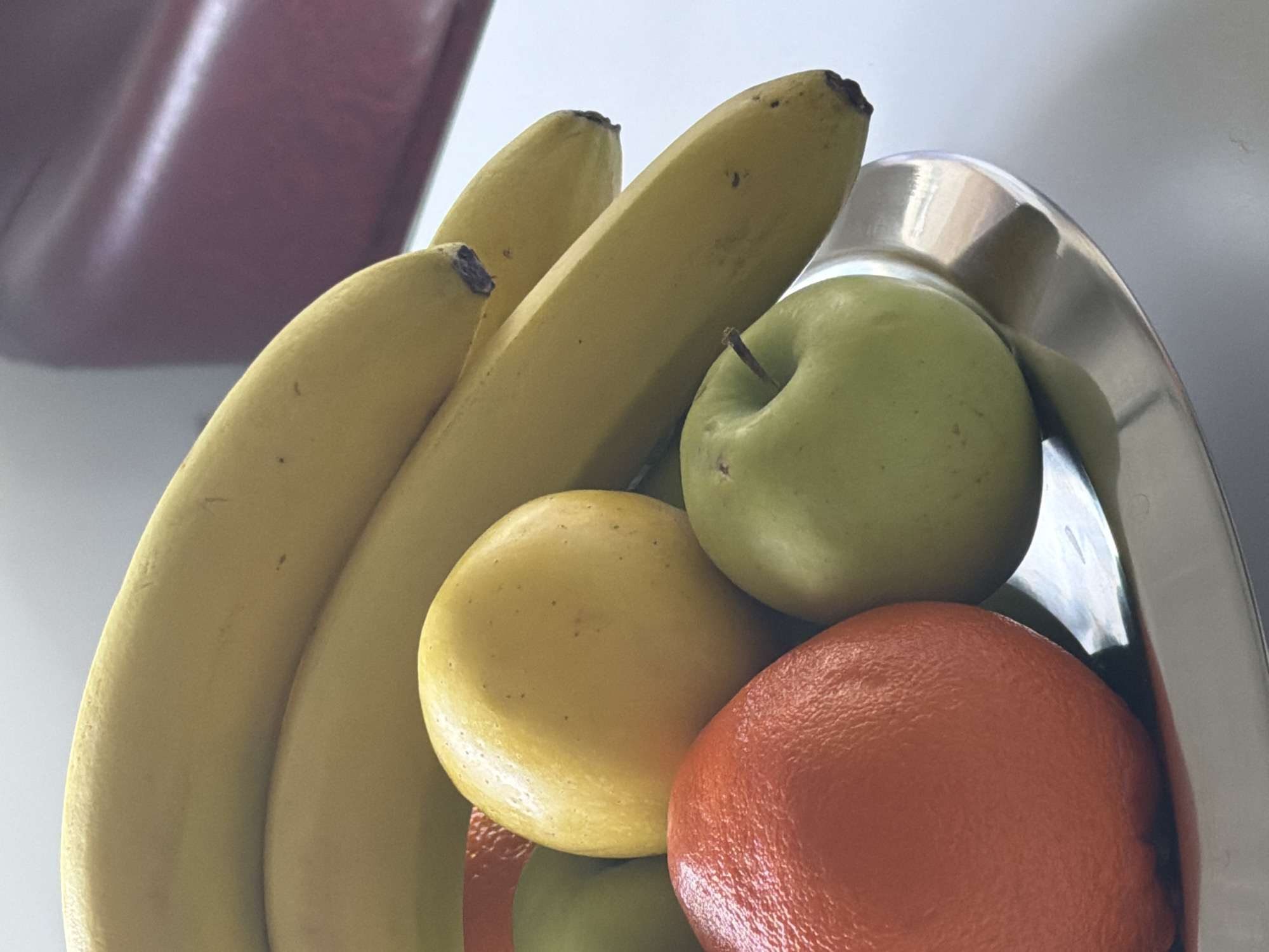 The image shows a close-up of a fruit bowl. The bowl, made of a silver or metallic material, is filled with various fruits. There are three yellow bananas, a green apple, an orange, and a yellow lemon. Part of a red object is also seen in the background. There is no indication that the image was taken in Yerevan, Armenia.