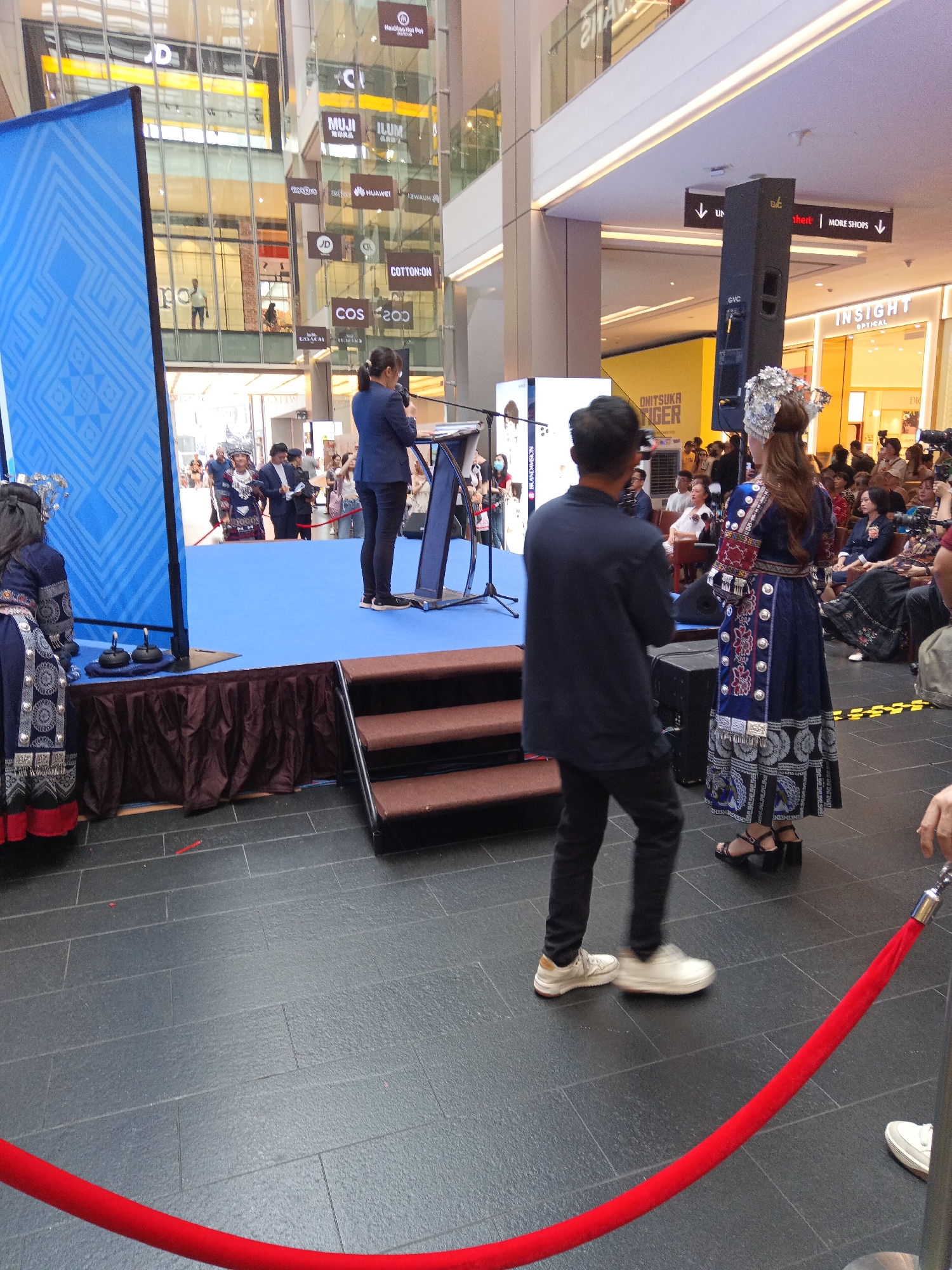 The image depicts an indoor event, likely a cultural or promotional gathering, held at the Sunway Pyramid Mall in Bandar Sunway, Petaling Jaya, Selangor, Malaysia, which is part of the Greater Kuala Lumpur area. A stage is set up with a blue backdrop featuring a geometric pattern. A woman in a dark suit stands at a podium on the stage, appearing to be speaking. To her right, two women in traditional ethnic attire are also on the stage, one wearing an elaborate headdress and the other in a decorated blue dress.
In the foreground, a man in a dark jacket and black pants stands facing away from the camera, looking towards the stage. Another person, whose lower legs are visible, is also in the foreground, wearing white shoes. A red velvet rope barrier is positioned across the lower part of the image, suggesting a designated viewing area.
The background reveals the multi-level interior of a modern shopping mall. Store signs are visible on the upper floors, including "MUJI," "COS," "COTTON-ON," and "INSIGHT OPTICAL." People are scattered throughout the mall, some watching the event and others browsing. The overall atmosphere appears to be that of a public event within a commercial setting.