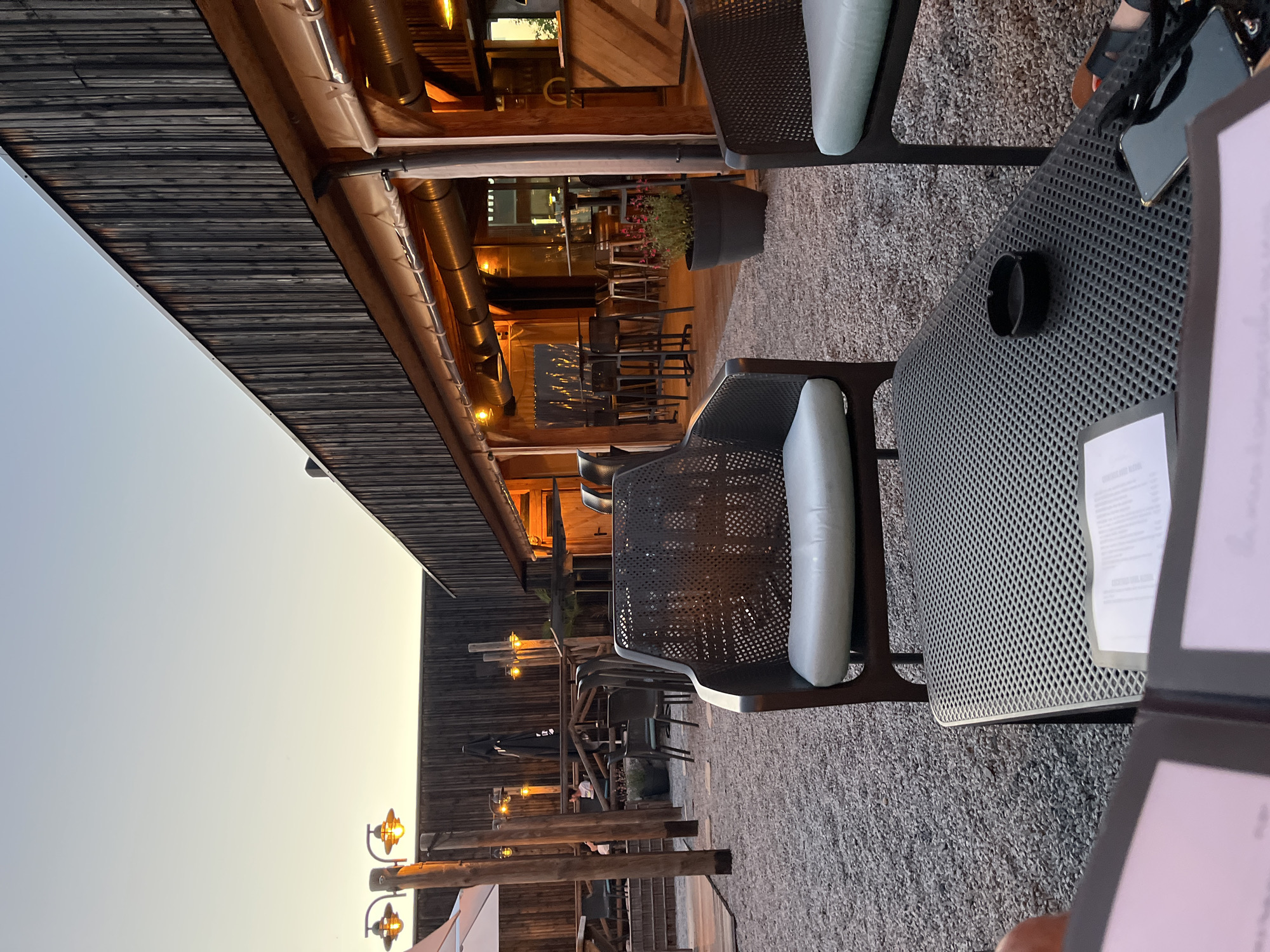 The image shows an outdoor seating area at a restaurant or bar in Andrézieux-Bouthéon, France. The area has metal tables and chairs with light-colored cushions on a gravel surface. Some tables have ashtrays, menus, and personal items. The setting sun indicates the image was taken during the evening. The building structure includes dark wooden walls, beams with string lights, and exposed ventilation systems. Several people are seated at other tables in the background, along the railing.