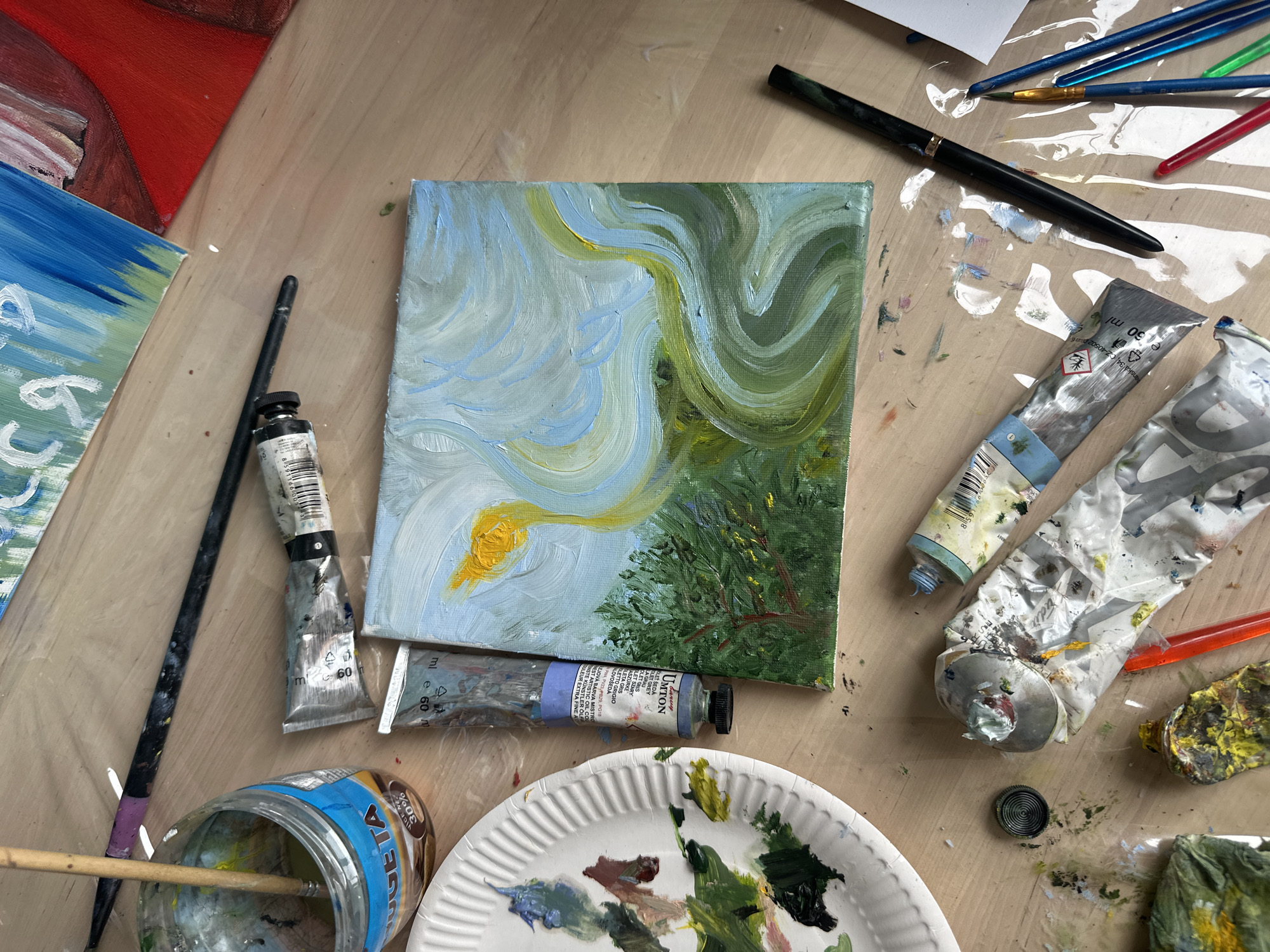 The image shows an overhead view of an artist's workspace. The primary subject is a small, abstract oil painting depicting swirling patterns of green and blue with a yellow element, likely representing a bird or a stylized form. The painting is placed on a wooden surface, surrounded by art supplies. These include several tubes of oil paint, some of which are partially squeezed and splattered with color. Visible text on the paint tubes indicates brands like "UMTON" and "Winsor & Newton," with "60 ml" indicating the volume. Various paintbrushes are scattered around, including one with a purple tip and another with a wooden handle partially submerged in a jar of what appears to be water or solvent. The jar has a label with "Velteta" and "30% Gratis" text. A white paper plate is being used as a palette, with dabs of oil paint in various shades of green, brown, and blue. There are also other unfinished canvases in the background, one with red and white abstract shapes, and another with blue and green strokes. The scene suggests an ongoing art project, with the lighting appearing to be artificial or from an overcast day, given the diffused light and lack of strong shadows. The overall impression is one of creative activity and a somewhat messy, but organized, studio environment.