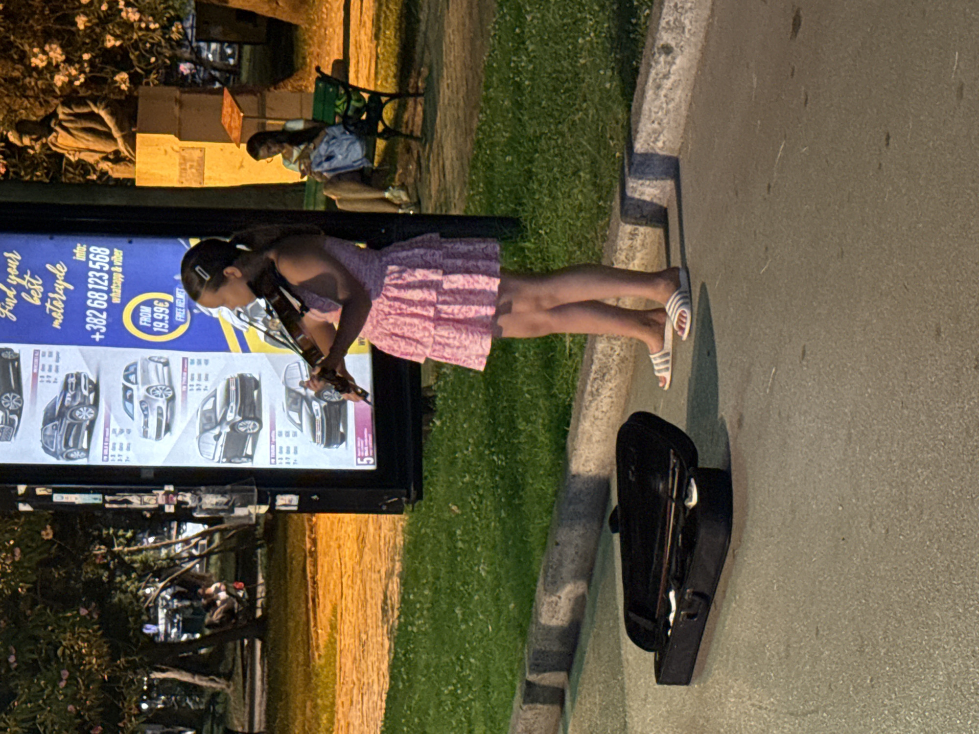 A young girl in a pink dress plays the violin on a sidewalk in Budva, Montenegro. Her violin case, open, lies on the asphalt in front of her. Behind her, a billboard advertises motorcycle rentals. Further back, a bronze statue stands on a pedestal, and a woman sits on a park bench, looking at her phone. The scene is illuminated by artificial lights, suggesting it is nighttime or evening. The grass and trees in the background are partially visible.