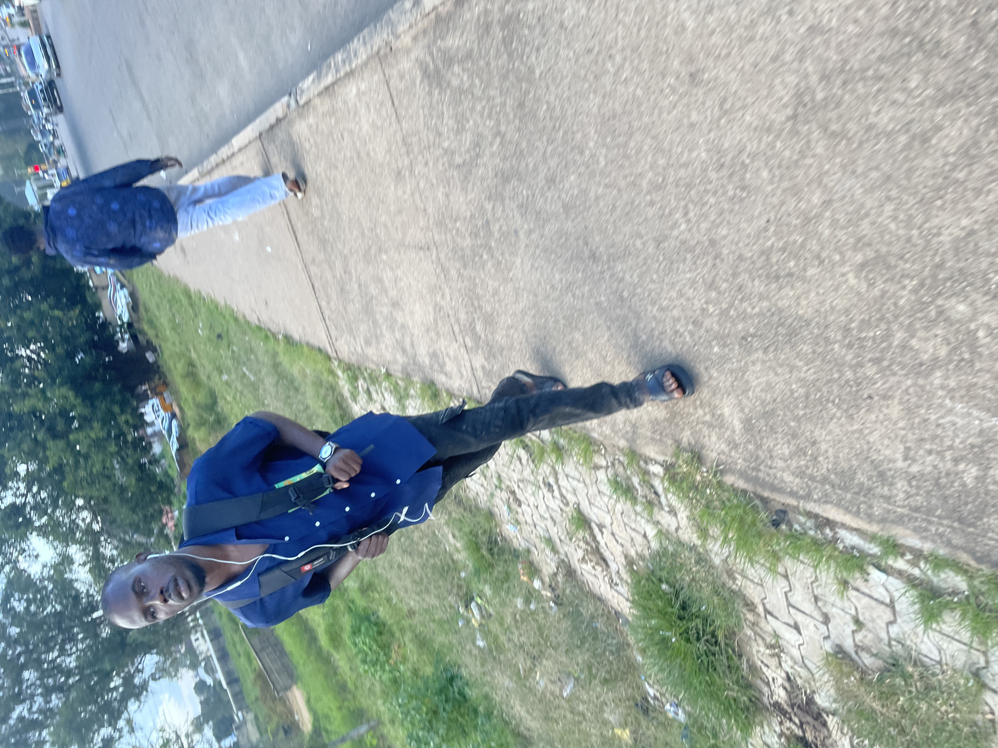 Two young men are walking on a paved sidewalk next to a road in Gura Top, Nigeria. The man in the foreground, wearing a blue short-sleeved shirt, black pants, and black sandals, is walking towards the right and appears to be in motion, possibly running or jogging, with a black backpack on. He has white earbuds in his ears and a watch on his left wrist. The second man, in the background, is walking away from the camera, wearing a dark blue patterned shirt and light blue pants. The environment shows a grassy area with some litter to the left of the sidewalk, and a road with several vehicles in motion to the right. Trees and buildings are visible in the distance, suggesting an urban or semi-urban setting.