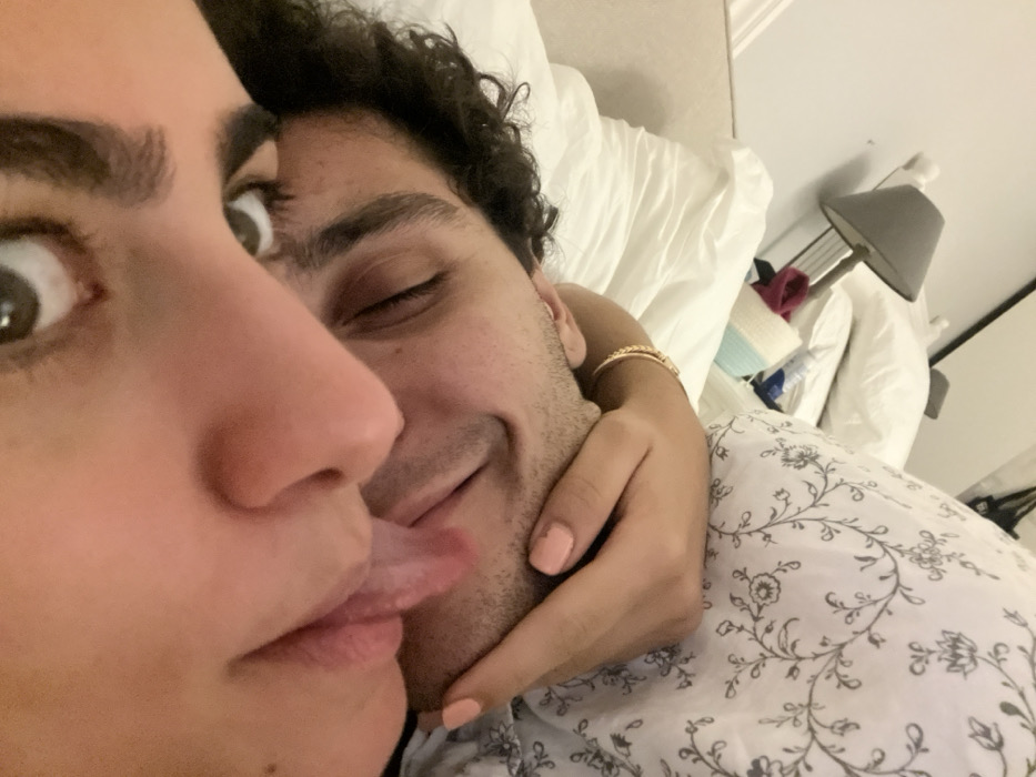 A close-up shot shows two people in bed. The person in the foreground has wide eyes and is sticking their tongue out. The other person, slightly behind, has closed eyes and a smile. Their hand is holding the face of the person in front. The bed has white pillows and a patterned sheet. A lamp is visible in the background, along with other miscellaneous items on a bedside table.