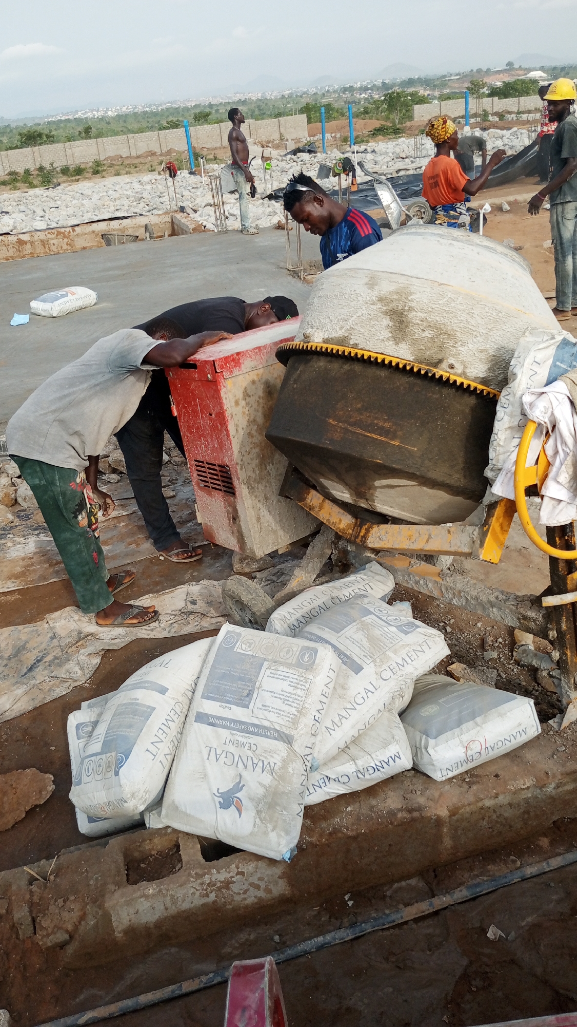 The image shows a construction site with several workers and equipment. One worker is leaning on a red power generator, while another is standing near a cement mixer. Several bags of "MANGAL CEMENT" are stacked nearby. Other workers are scattered throughout the site, and the background shows a cityscape. The exact location is unknown.