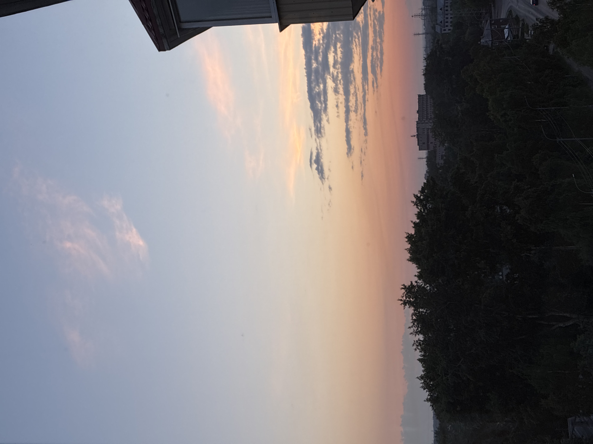 The image shows a sunset view from an elevated position, likely a balcony or window of a building. The sky transitions from a pale blue at the top to hues of pink and orange near the horizon. A few wispy clouds are visible in the upper part of the sky, with more textured, elongated clouds closer to the horizon. The foreground is dominated by the dark silhouettes of trees. Beyond the trees, a cityscape is visible, with buildings of various sizes and architectural styles, including what appears to be a large industrial or institutional building with multiple windows and a light-colored facade. Power lines and streetlights are also present, suggesting an urban environment. A road with a vehicle is visible in the lower right portion of the scene. The overall impression is of a late evening in an urban area, with the sky painted by the setting sun.