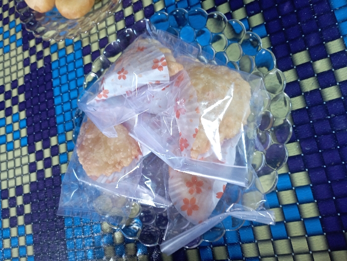 The image captures a close-up view of several individually packaged food items displayed on a clear glass plate, which rests on a vibrant, patterned mat. The primary subjects are small, roundish, light-brown pastries or sweets, each carefully placed inside a white paper cupcake liner. These liners feature a delicate pattern of small, red or orange floral designs, resembling cherry blossoms. Subsequently, each pastry within its liner is sealed inside a clear plastic bag. At least four such packaged items are visible, clustered on the main glass plate.
The glass plate itself is a notable object, characterized by its clear material and a distinctive scalloped or bubble-textured rim. In the upper left background, a portion of a second clear glass plate is visible, holding other food items that appear to be spherical and light brown, similar in color to the main pastries but without individual packaging.
The setting is an indoor space, likely a table or countertop, covered by a mat or tablecloth. This mat features a striking checkered pattern composed of squares in varying shades of blue (light blue, medium blue, dark blue) and a contrasting light yellow or light green. There are no people visible in the image, and no discernible text appears on any of the packaging or food items. The scene suggests food prepared for a gathering, sale, or personal consumption, with the individual packaging hinting at ease of distribution or a curated presentation. The location context for this scene is Pubian, Indonesia.