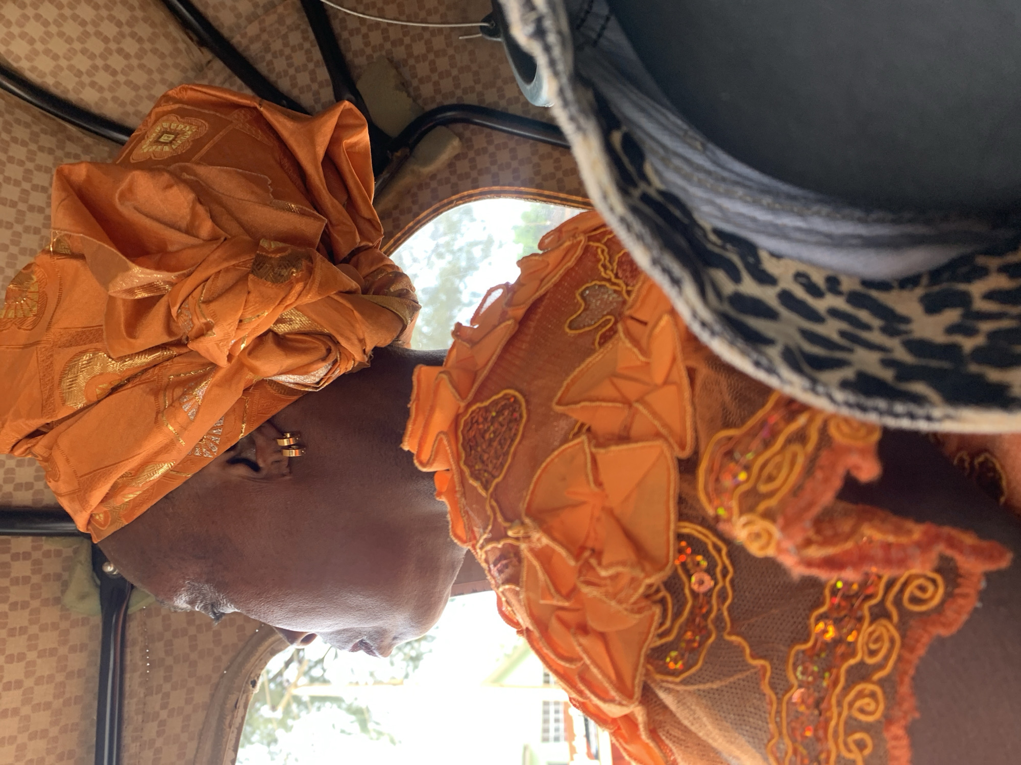 An adult individual, observed in a side profile, is seated inside a vehicle in Kabong, Nigeria, looking out a window to the left. The person has dark skin and wears a voluminous, orange headwrap (gele) embellished with gold patterns. The visible ear features two small, gold hoop earrings. The individual's attire includes an orange garment with a detailed neckline/shoulder area, characterized by ruffled fabric, intricate gold embroidery, and iridescent sequins on a sheer, mesh-like overlay.
The vehicle's interior features a light brown, checkered-patterned ceiling and black metal support bars. Through the blurred window, bright daylight illuminates green foliage and a distant, multi-story white building with windows. In the foreground on the right, a portion of an object with a black and tan leopard print pattern is visible, adjacent to a dark, indistinct object. Two thin wires or strings descend from the ceiling towards the right side of the frame.
