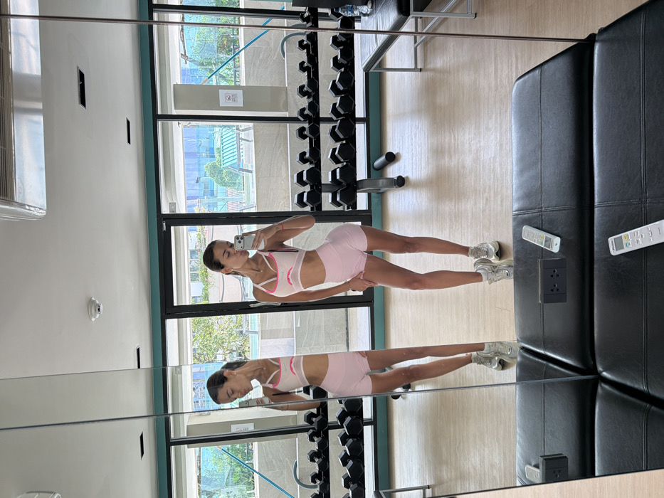 A young woman in a pink sports bra and shorts takes a mirror selfie in a gym. Weights line the wall near a window overlooking an outdoor area. The mirror reflects her image, showing a symmetrical view of her and the gym equipment. To the right of the mirror, a black leather couch is visible with a pair of remote controls.