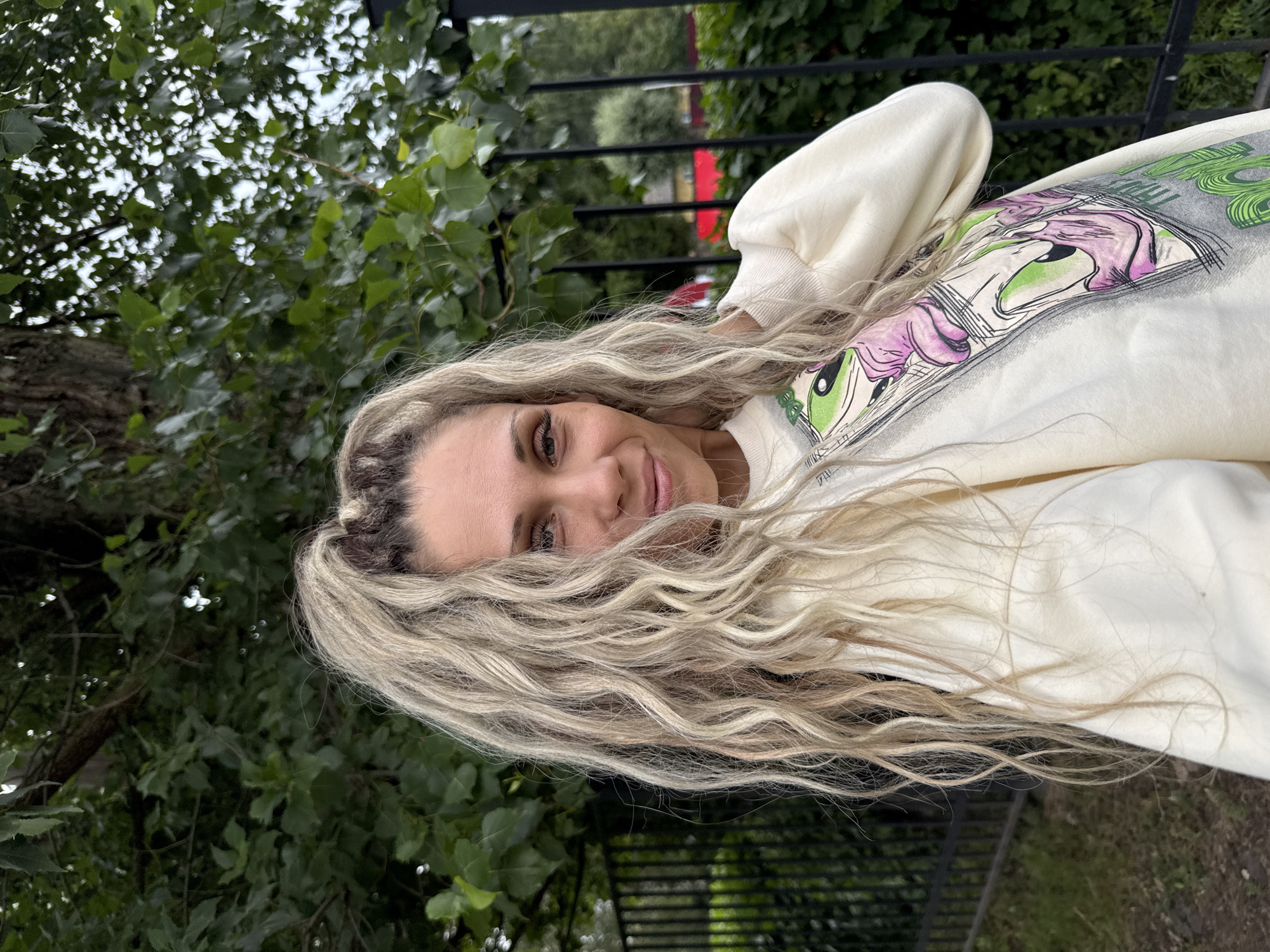 A woman is pictured from the chest up, outdoors in a partially shaded environment. She has long, blonde, curly hair pulled back into a high ponytail, with some strands framing her face. Her eyes are looking towards the camera, and she has a slight smile. She is wearing a cream-colored sweatshirt with a graphic print featuring cartoon characters. Behind her, there is a black metal fence, green foliage, and the trunk of a large tree. The background is slightly out of focus, suggesting a shallow depth of field. The location is identified as Rusanovo, Russia.