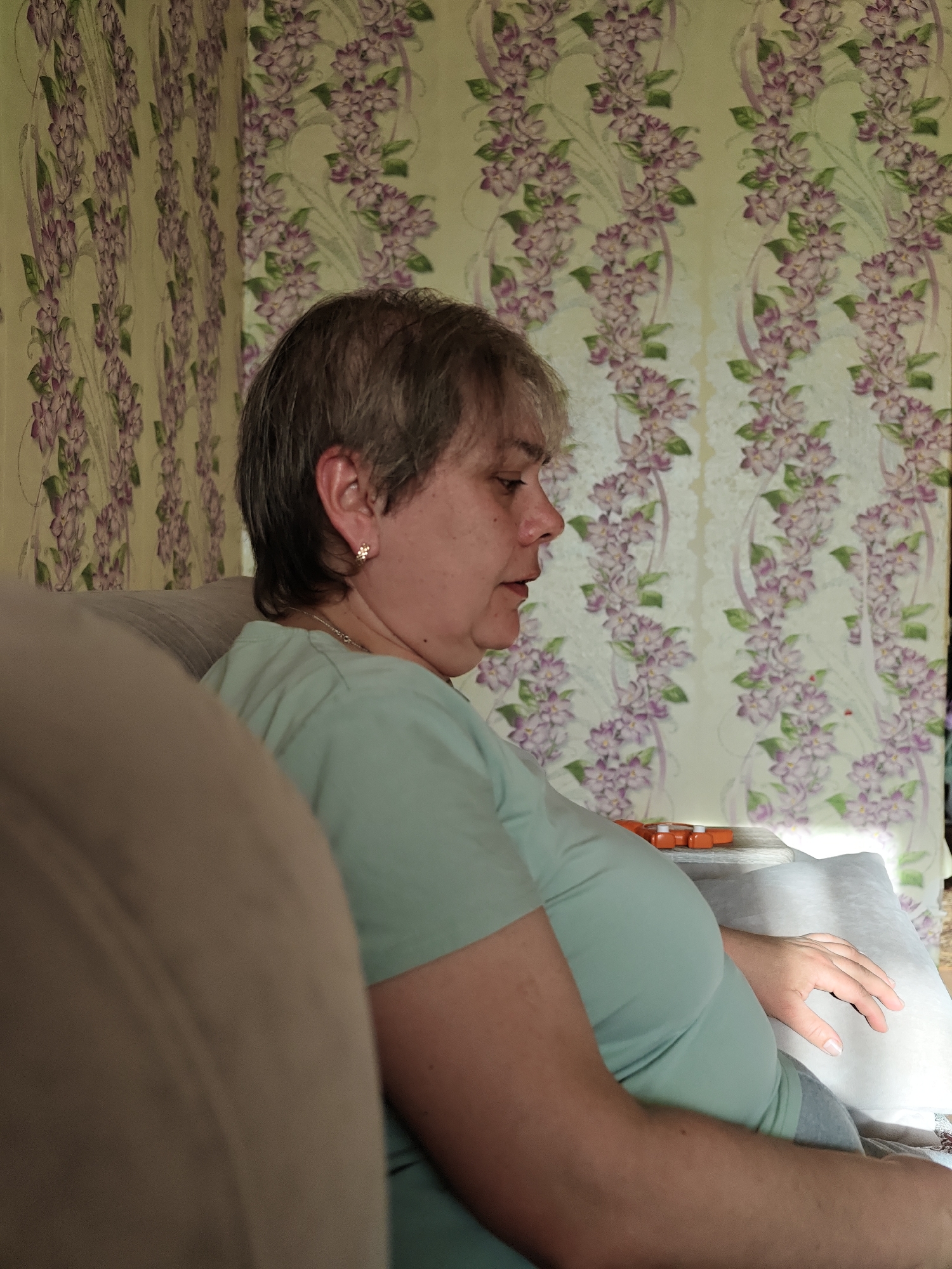 A middle-aged woman with short, gray hair sits on a couch. She is wearing a light green t-shirt and has a small earring in her left ear. She is looking to her right. Her right hand rests on a gray pillow. The couch is beige. Behind her, the wall is covered in floral wallpaper with purple and white flowers. There is also a package of orange medicine on a table behind the woman. The location is Уральское, Russia.
