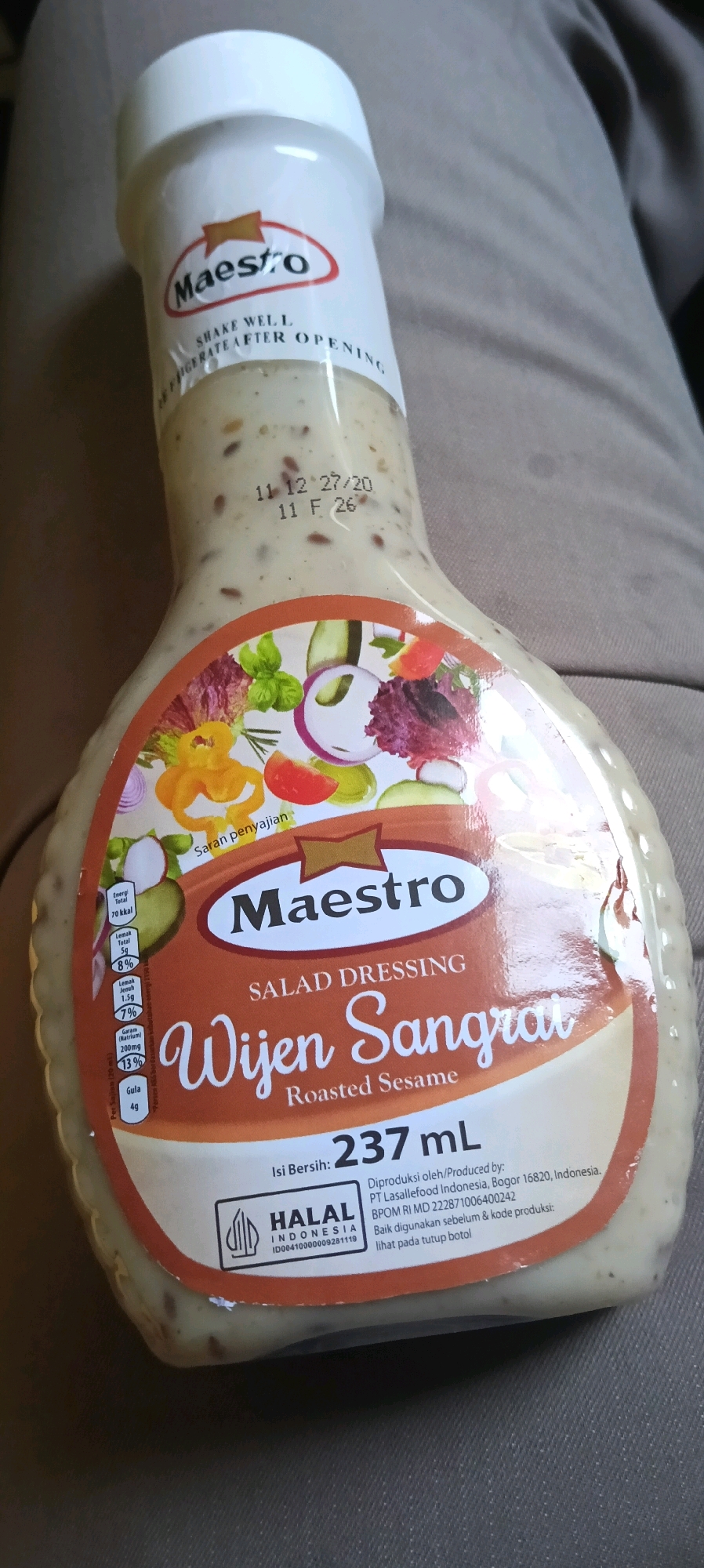The image shows a close-up of a bottle of Maestro Roasted Sesame Salad Dressing. The bottle has a white cap and a label with a stylized illustration of salad ingredients such as lettuce, tomatoes, onions, and cucumbers. The brand name "Maestro" is prominently displayed in a red oval with a gold starburst. Below this, "SALAD DRESSING" is written, followed by the product name in Indonesian, "Wijen Sangrai," and its English translation, "Roasted Sesame." Nutritional information, including serving size and calorie count, is visible on the left side of the label. A "HALAL INDONESIA" certification is also present. The expiration date "11 12 27/20" and batch code "11 F 26" are stamped on the bottle. The bottle appears to be slightly tilted and is resting on a textured surface, possibly fabric. The background is blurred and shows a muted, neutral color. There are no discernible elements within the image that would indicate a specific location in Kuala Kapuas, Indonesia, or any interaction with people.