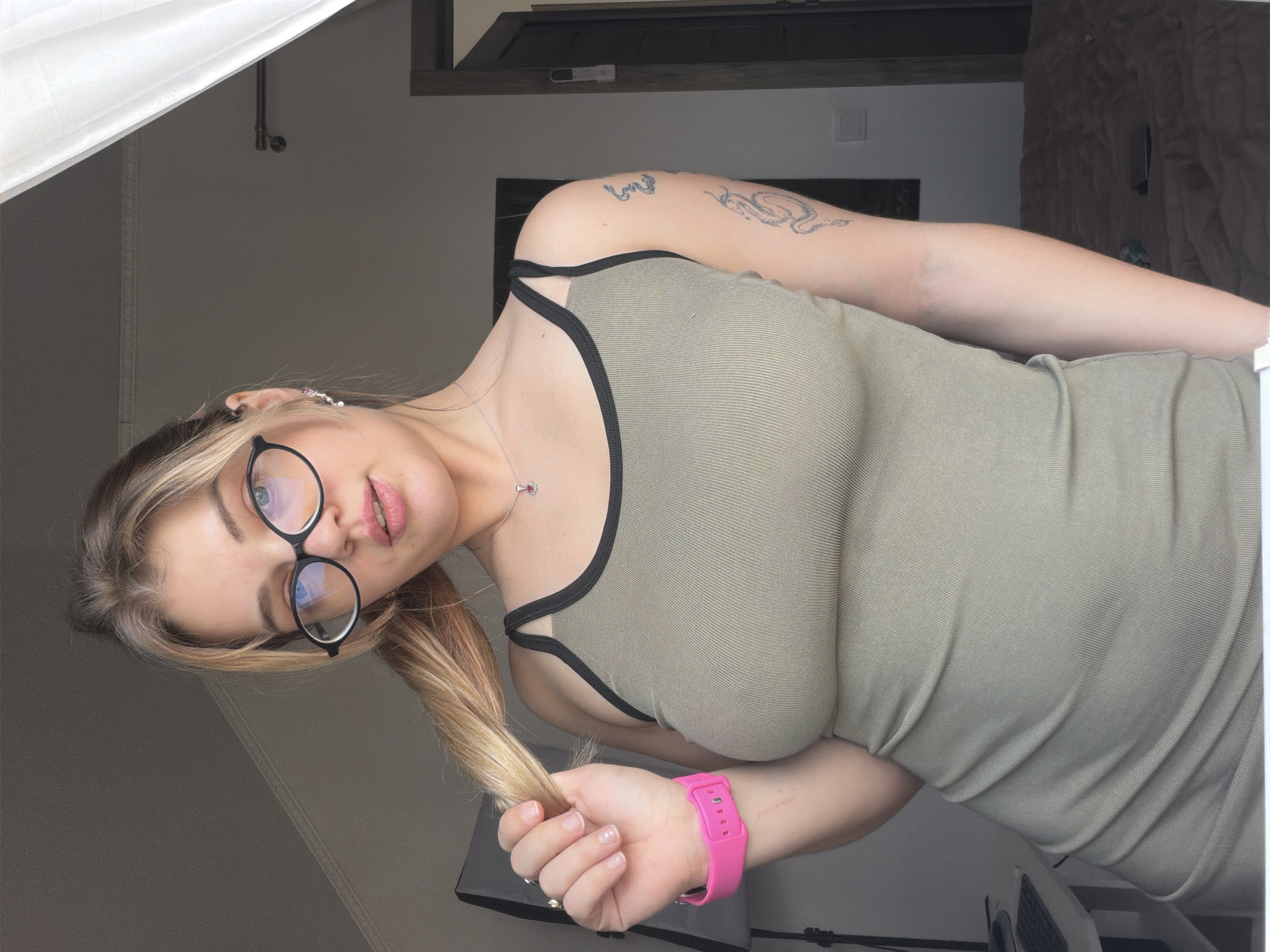 The image shows a woman wearing glasses and a green ribbed tank top with black straps. She has blonde hair and fair skin with tattoos on her left arm. She is holding a portion of her hair in her right hand, which has a pink smartwatch on the wrist. The background includes a partial view of a room with gray walls, a white curtain, and what appears to be a shelf or cabinet above. While it's impossible to confirm with certainty from the image, it can not be determined that the photo was taken in Большая Шильна, Russia.