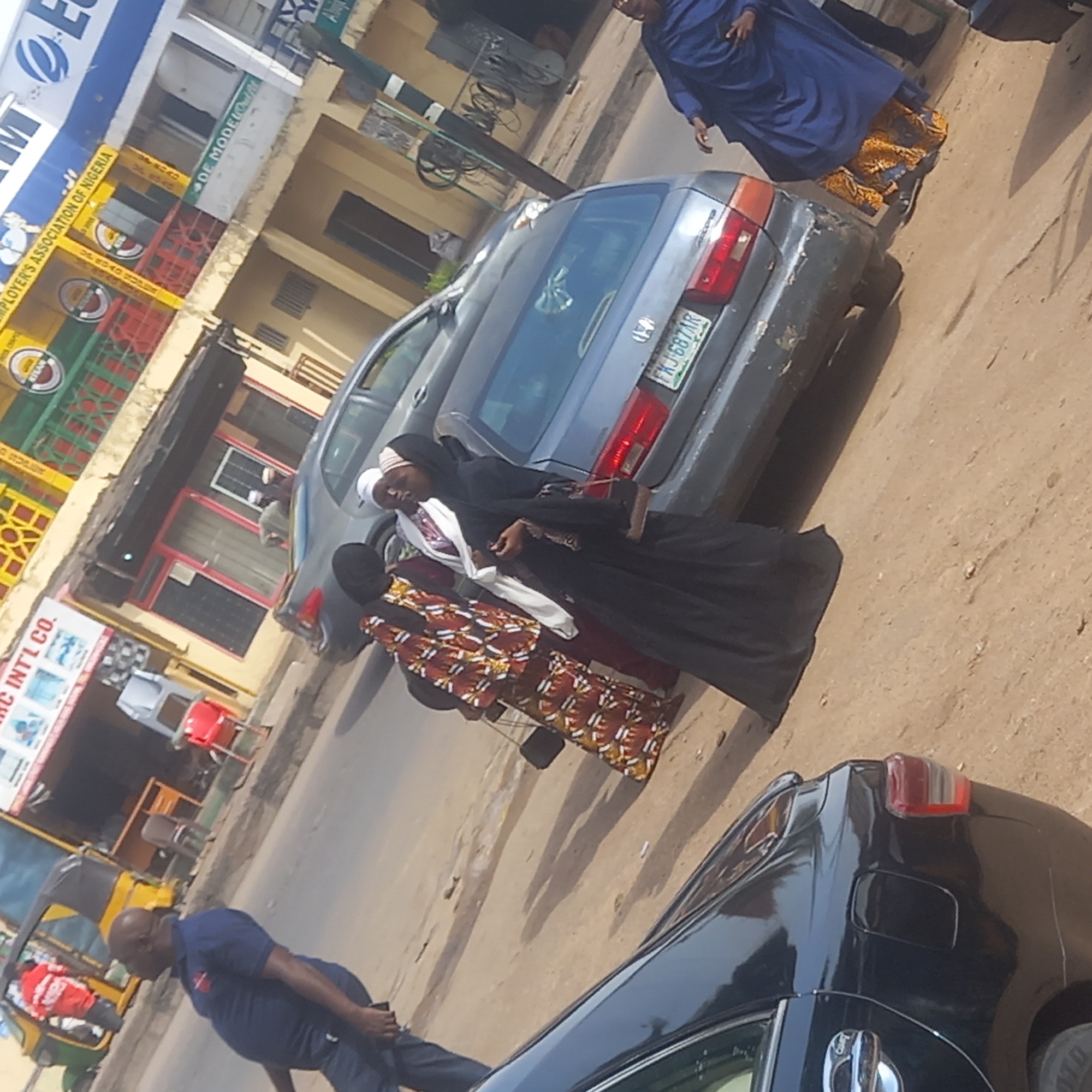 The image shows a street scene in Dadi Riba, Nigeria. Three women wearing hijab head coverings and long dresses are walking on the street next to two parked cars. A man in a blue polo shirt and dark pants is walking in the opposite direction. A yellow and green vehicle, possibly a rickshaw, is visible in the background. Buildings with signs, including one that reads "RC INT'L CO." and another that includes "EMPLOYER'S ASSOCIATION OF NIGERIA," are along the side of the street. The license plate on one of the cars reads "FKJ-687AR."