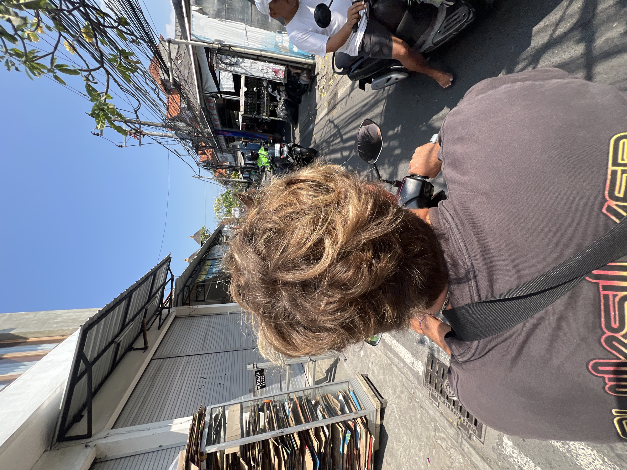 The image depicts a first-person perspective of someone riding a scooter down a street in Kuta, Indonesia. The rider's sun-bleached, wavy hair is in the foreground. They are wearing a dark t-shirt with a logo on the back. A black strap is visible across their shoulder. To the left of the rider, a tall, narrow metal rack is stacked with cardboard boxes. The street is lined with buildings, many with closed metal roll-up doors, suggesting a commercial area. Overhead, a dense network of electrical wires crisscrosses the sky, a common sight in Indonesian cities. Further down the street, other scooters and motorcycles are visible, with people riding them. One man in a white t-shirt and shorts is on a scooter to the right, with his bare feet visible. The sky is clear and blue, indicating a sunny day. The overall scene is characteristic of a bustling street in a Southeast Asian city.