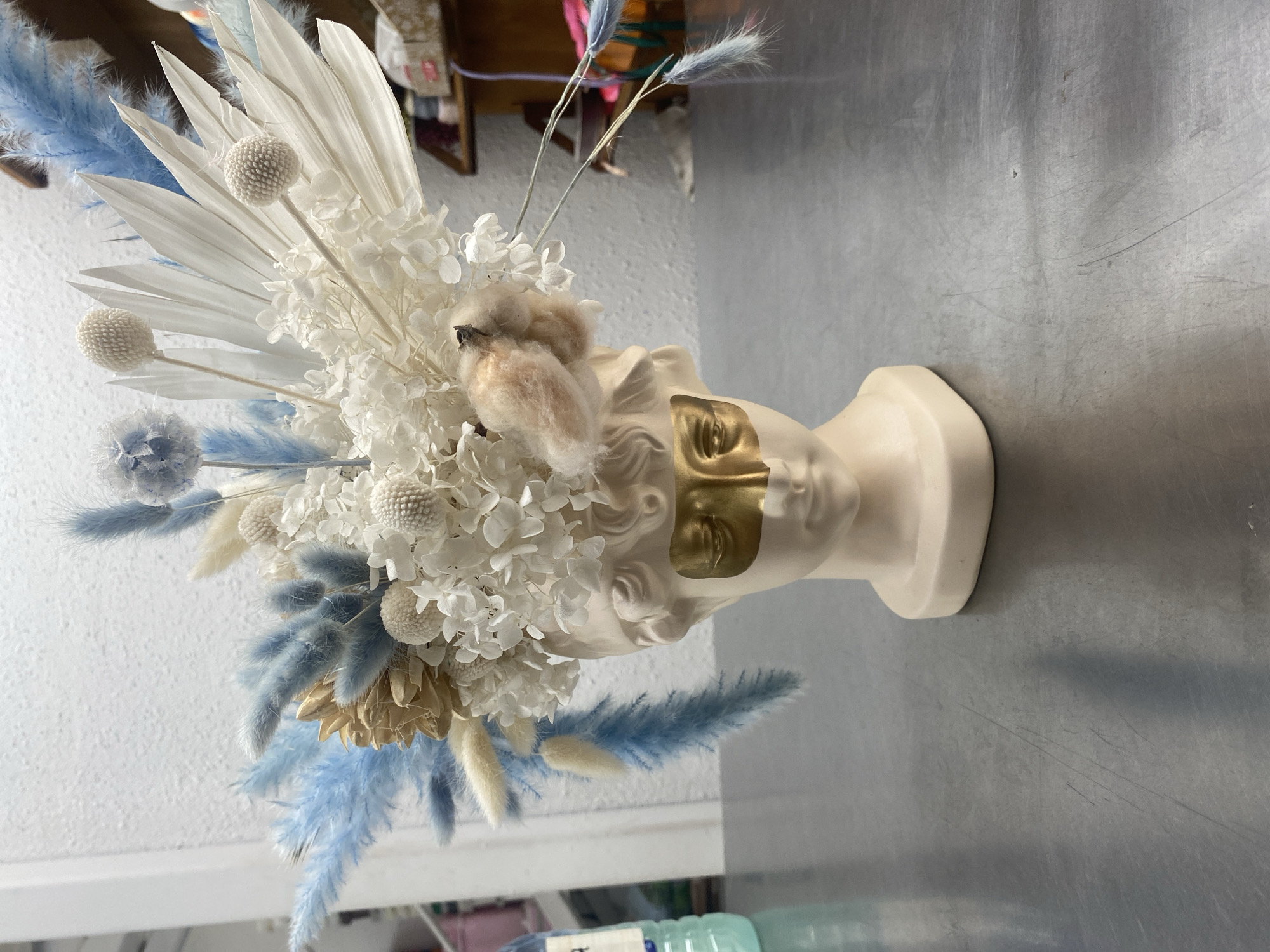 The image displays a floral arrangement in a light beige bust-shaped vase with gold leaf accents on the eyes. The arrangement features dried flowers in a palette of white, cream, and light blue. The blooms include hydrangea, cotton, and various types of ornamental grasses. The vase rests on a gray metallic surface. The background suggests an interior setting, with hints of furniture and other objects. The image was not taken in Yekaterinburg.