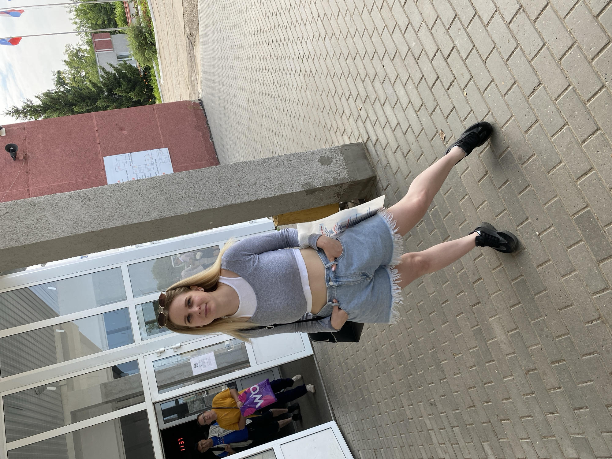 A young woman with blonde hair and sunglasses on her head stands in front of a building in Chaikovsky, Russia. She is wearing a grey long-sleeved crop top, a white undershirt, and denim shorts with frayed edges. She has her hands in her pockets and is looking towards the camera. In the background, two men and a woman are entering or exiting the building through glass doors. The building appears to be a public facility, possibly a train station or administrative building, with a red facade and large windows. The ground is paved with grey interlocking bricks. The sky is overcast.