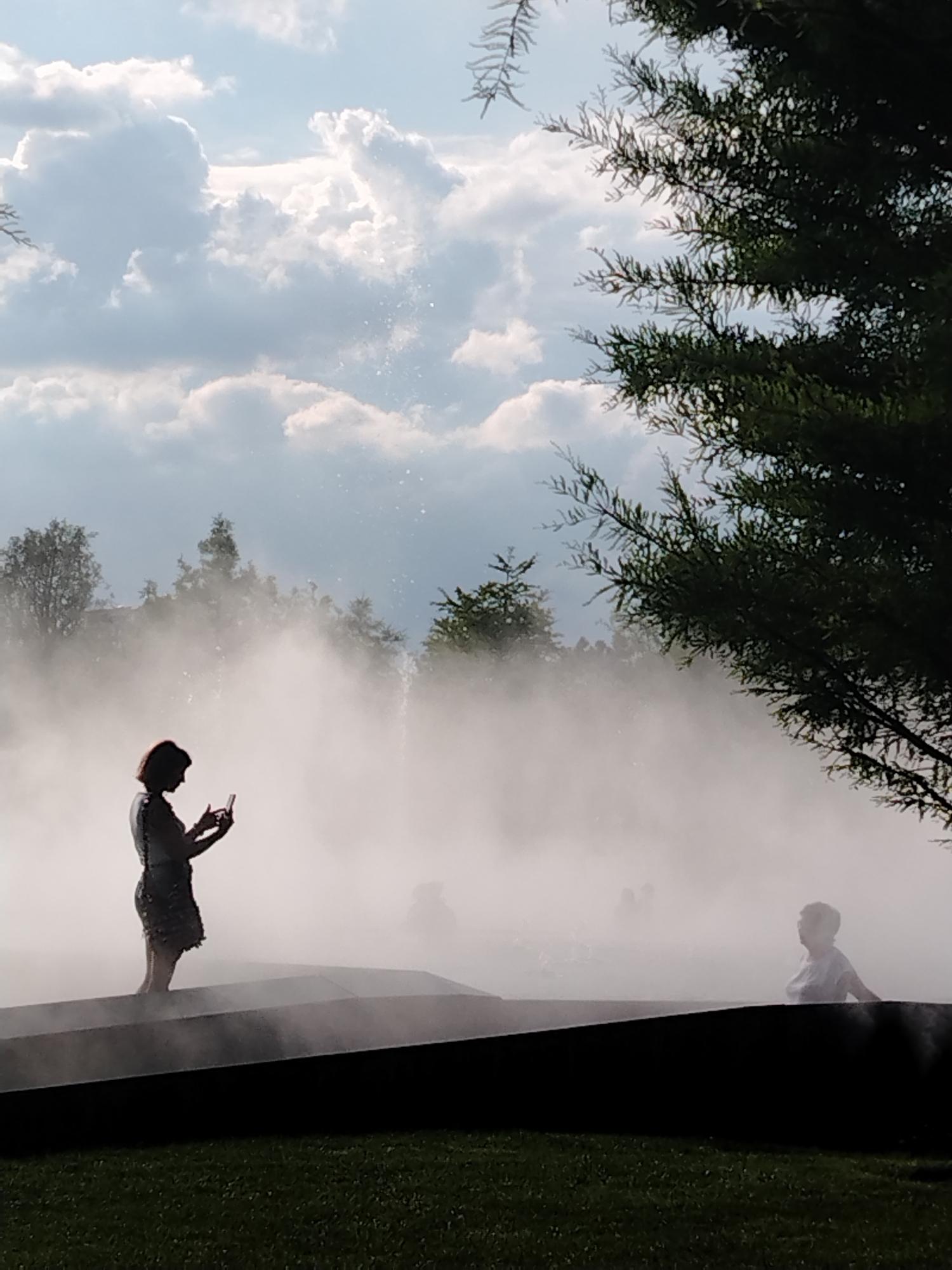 The image depicts a public park with a water mist installation, likely in Krasnodar, Russia, given the context. In the foreground, a person, silhouetted against the mist and sky, stands on a dark, angular platform, holding a mobile phone and appearing to take a picture or record a video. To the right, another silhouetted figure, possibly a child, is visible in the distance, also within the mist. The background features trees and a sky filled with dramatic, light-colored clouds. The mist creates a hazy, atmospheric effect, obscuring some details of the environment and other individuals who may be present further back. The overall scene suggests a leisurely outdoor activity in a park setting.