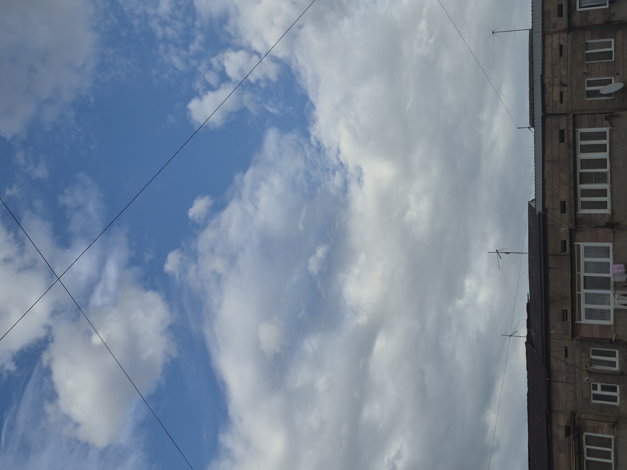 The image, oriented correctly, presents an upward view of an urban scene, divided between a sky on the left and a building on the right. The sky is partially clear blue with scattered white, wispy clouds, while a significant portion, especially towards the right, is covered by larger masses of greyish-white clouds. Two thin, dark lines, likely power or communication cables, diagonally cross the sky from the upper left to the lower right. Another horizontal cable is visible lower down.
On the right, a section of an apartment building is visible. It appears to be an older, multi-story structure with a textured, brownish-grey facade made of stone or concrete blocks. The building features several windows with white frames and multiple balconies. A white satellite dish is affixed to one of the walls or balconies. Notably, pink clothing is hanging on a line outside an upper window or balcony, drying. The dark, flat roofline of the building is visible, equipped with several antennas, including one distinct starburst-shaped antenna, from which wires extend.
The scene suggests daytime with varied cloud cover. No people are visible, and no specific activity is occurring beyond clothes drying. The architectural style and details like the visible antennas and clothesline are consistent with residential areas in cities such as Yerevan, Armenia. There is no visible text in the image.