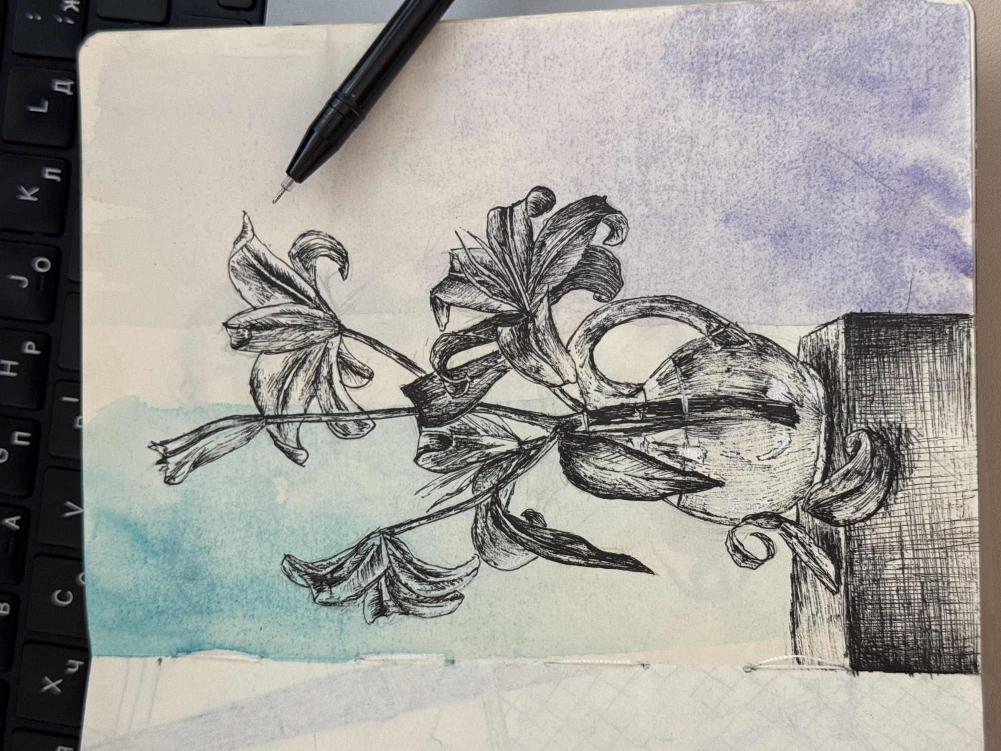 A sketchbook page features a pen-and-ink drawing of lilies in a vase. The drawing is set against a background of watercolor washes in shades of blue and purple. A black pen rests on the upper right corner of the page. The lilies are rendered with detailed linework, showing delicate petals and stems. The vase is depicted with a textured surface, and a handle is visible. Two fallen lily petals lie on the surface below the vase. The sketchbook page is open, resting on a dark surface, possibly a keyboard.