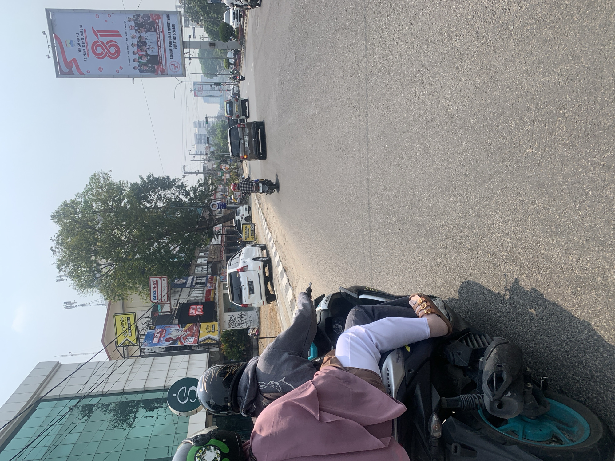 On a bright, sunny day in Palembang, Indonesia, two people are riding on a dark-colored scooter along a paved road. The rider, wearing a black helmet with a gold design, is in the front, and a passenger, wearing a pink hijab and white pants, is seated behind. They are positioned in the left lane of a multi-lane road. In the background, a billboard celebrating "DIRGAHAYU REPUBLIK INDONESIA 81" is visible. Other vehicles, including cars and motorcycles, are on the road, and buildings line the side. The scene suggests a typical urban street environment in Palembang.