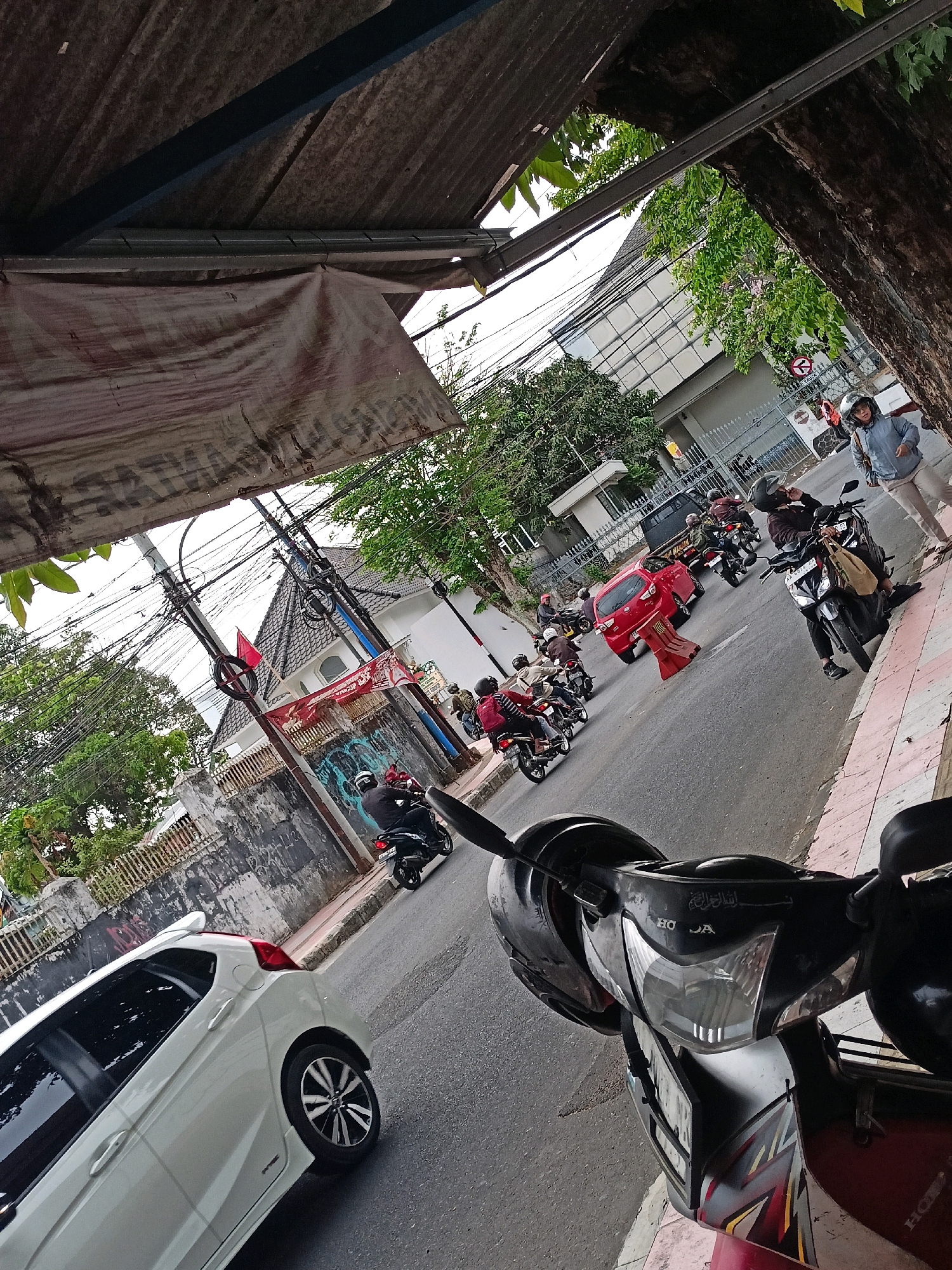 The image captures a street scene in Magelang, Indonesia, possibly during the daytime. A white car is parked on the left side of the road, its rear visible. The road itself has a moderate amount of traffic, consisting mainly of motorcycles and a red car. Several people on motorcycles are seen wearing helmets, with some carrying bags. A woman is standing on the sidewalk on the right, wearing a light blue jacket and beige pants, also with a helmet. A tree with green foliage is present on the right side of the frame, and electrical wires crisscross overhead. A building with a white facade and dark tiled roof is visible in the background, along with a fence. Graffiti is present on a wall. The scene appears to be an urban environment with typical street activity.