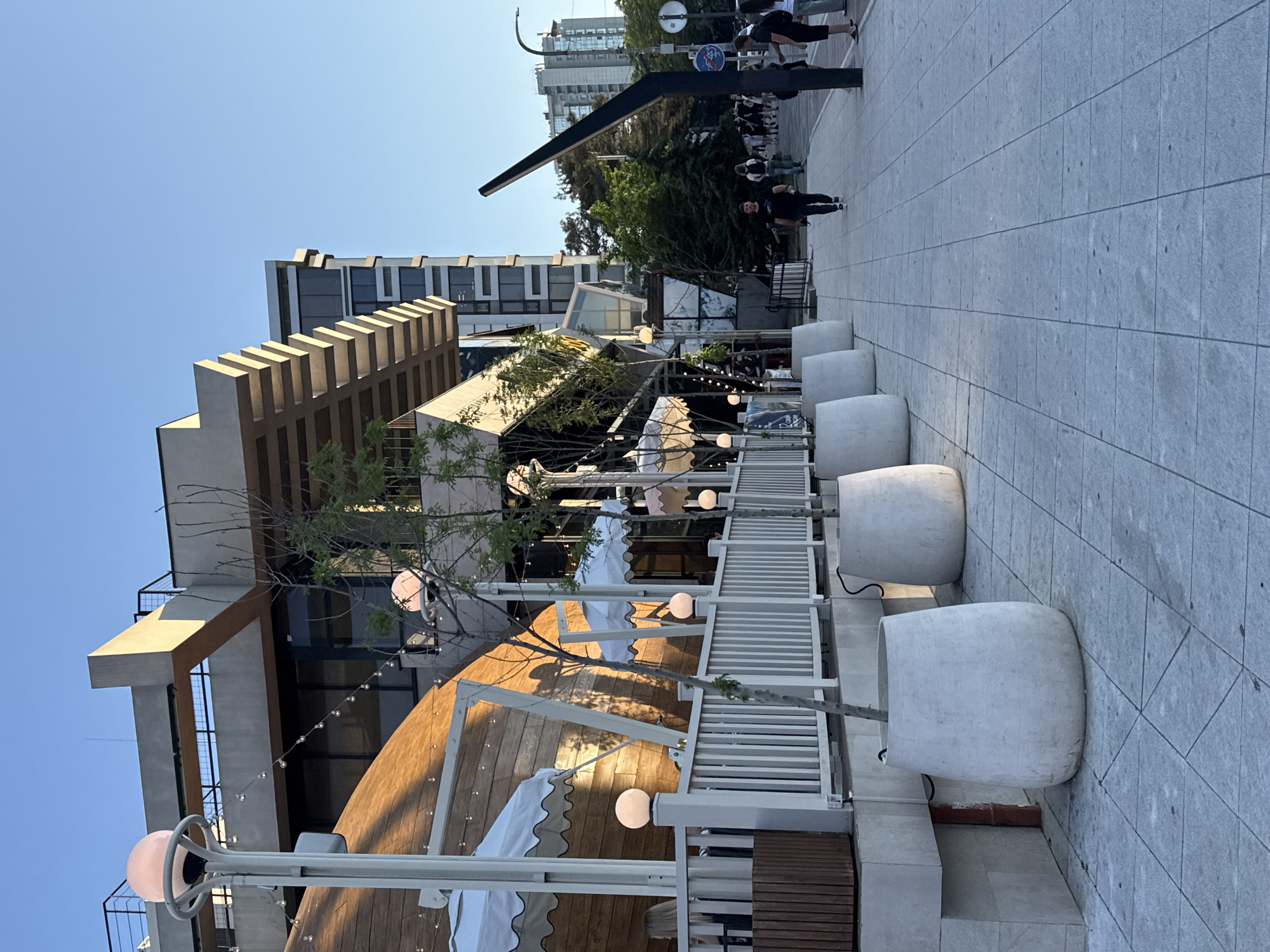 The image depicts a street scene in Neberdzhaevskaya, Russia, under a clear blue sky. The foreground features a paved sidewalk with a row of large, white planters containing trees. To the left of the sidewalk, a white fence separates it from an outdoor seating area with white umbrellas. Behind the fence, a curved wooden structure with windows and lights is visible, suggesting a restaurant or commercial establishment. Further back, a modern building with multiple stories and glass windows rises into the sky. Several people are walking on the sidewalk, some in groups and others individually. The overall impression is of a bustling urban environment with commercial activity and pedestrian traffic.