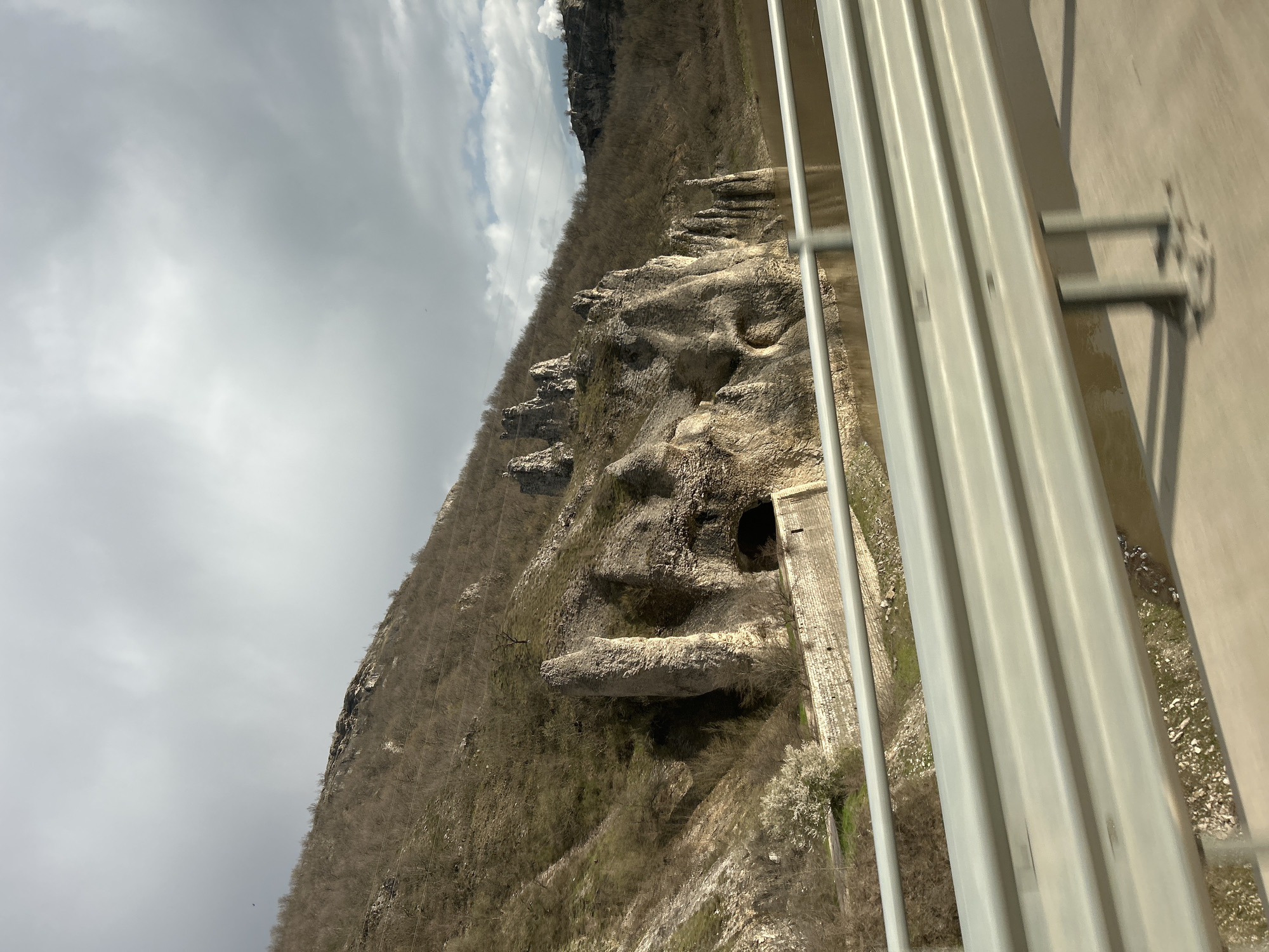 The image shows a rock formation resembling a human face on a mountainside in Asparuhovo, Bulgaria. The rock face is in profile, with a clearly defined forehead, nose, and chin. There appears to be a dark cave or opening that may represent an eye. The mountainside is covered in sparse vegetation. A metal guardrail is visible in the foreground, suggesting the photo was taken from a bridge or elevated platform. The sky is overcast.