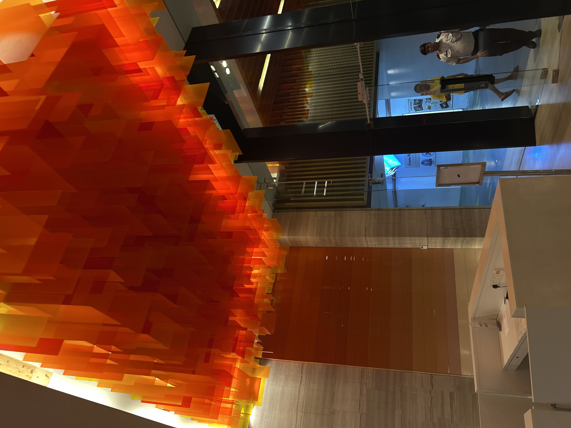 The photograph depicts the interior of a building in Kuala Lumpur, Malaysia. A large, abstract sculpture composed of numerous translucent orange and yellow rectangular elements dominates the left side of the frame, appearing to hang from the ceiling. To the right of the sculpture, a vertical strip of orange-colored wall panels is visible, adjacent to what appears to be a white structure with electrical outlets and possibly a small display. Further back, a glass partition or window reflects two people: one wearing a uniform, possibly a security guard, and another in a yellow shirt and shorts. The floor is light-colored, possibly made of marble or a similar material. The overall impression is of a modern, well-lit building with an artistic architectural design.