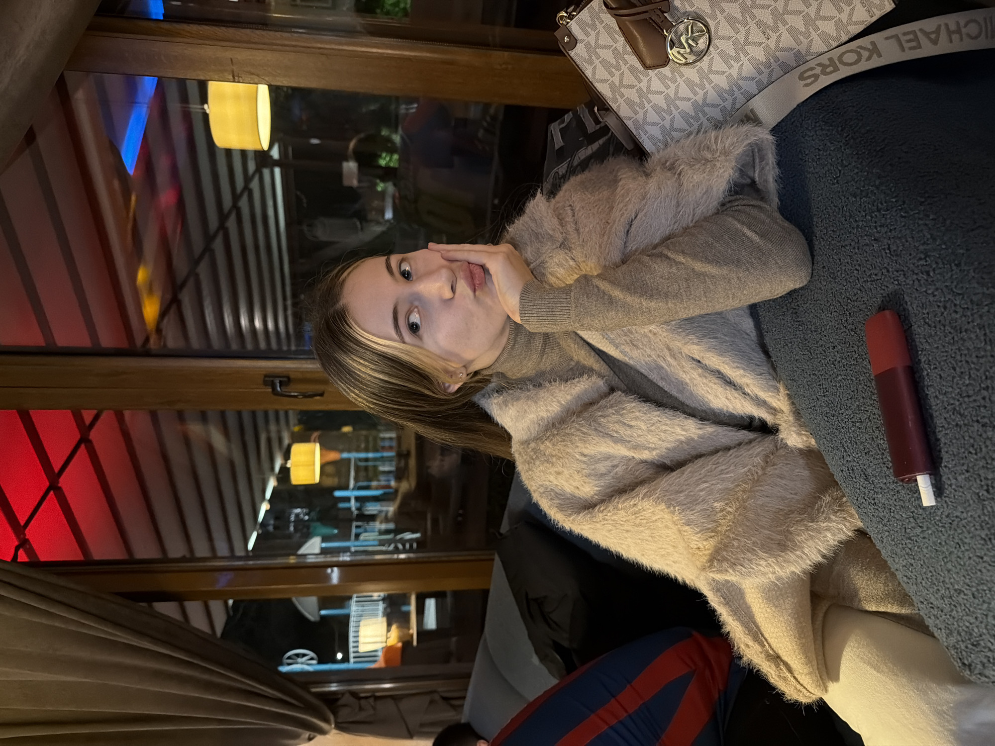 A young woman sits inside a restaurant. The environment appears to be Plovdiv, Bulgaria, due to the architectural style visible through the windows, including striped awnings. The woman is wearing a brown sweater and a fur-trimmed jacket. She has her hand on her face and is looking at the camera with a slight pout. Her hair is blonde with darker roots. Beside her is a Michael Kors purse. A small red object rests on a textured blue surface, presumably a seat. Outside the window, there are street lights and a view of the city. The light is dim inside, suggesting evening or nighttime. There is a heavy curtain on the bottom left corner.