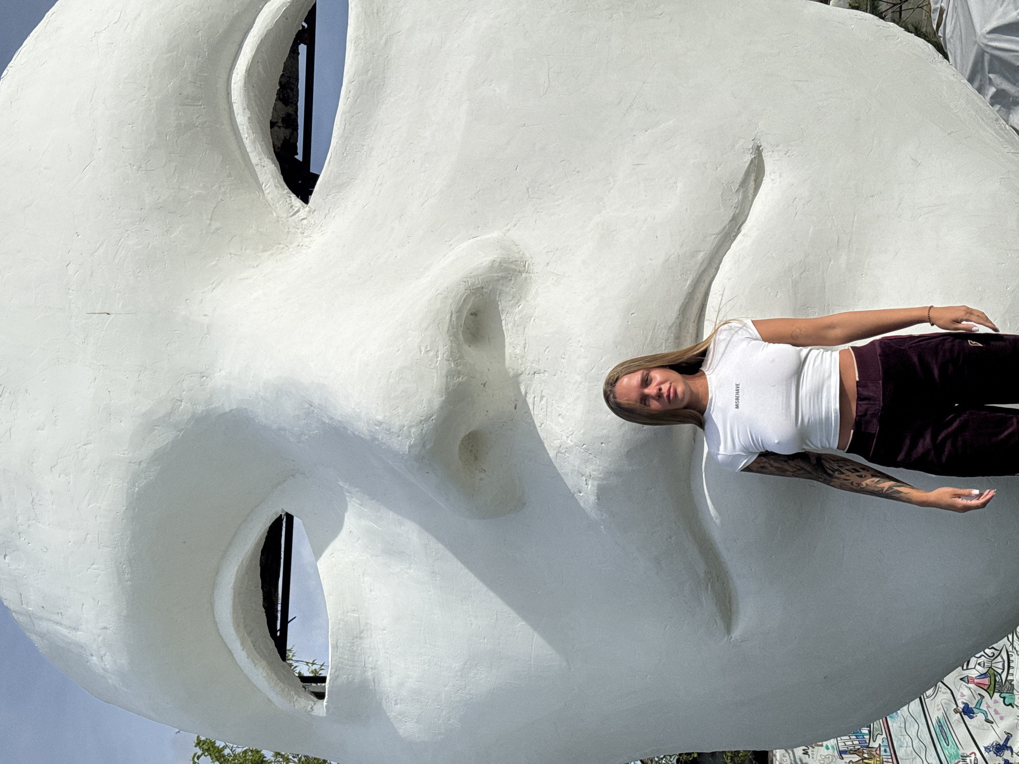 A person is photographed next to a giant concrete art installation of a face. The face is white, and the photograph is taken on a sunny day. The face installation has holes for the eyes. The location is in the city of Nizhny Novgorod, Russia.