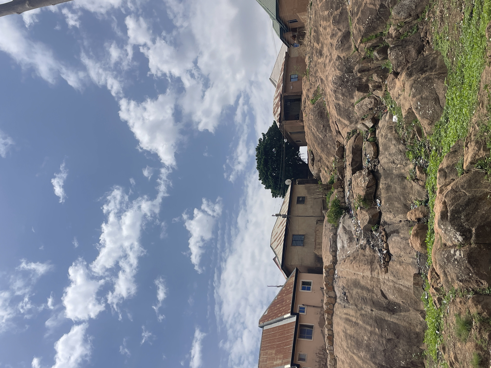 The image captures a scene in Jos, Nigeria, featuring buildings constructed on the side of a rocky outcrop. The houses are multi-storied, with visible windows and corrugated metal roofs. A tree is nestled among the buildings. The rocky terrain is interspersed with vegetation and some visible litter. The sky is blue with scattered clouds.