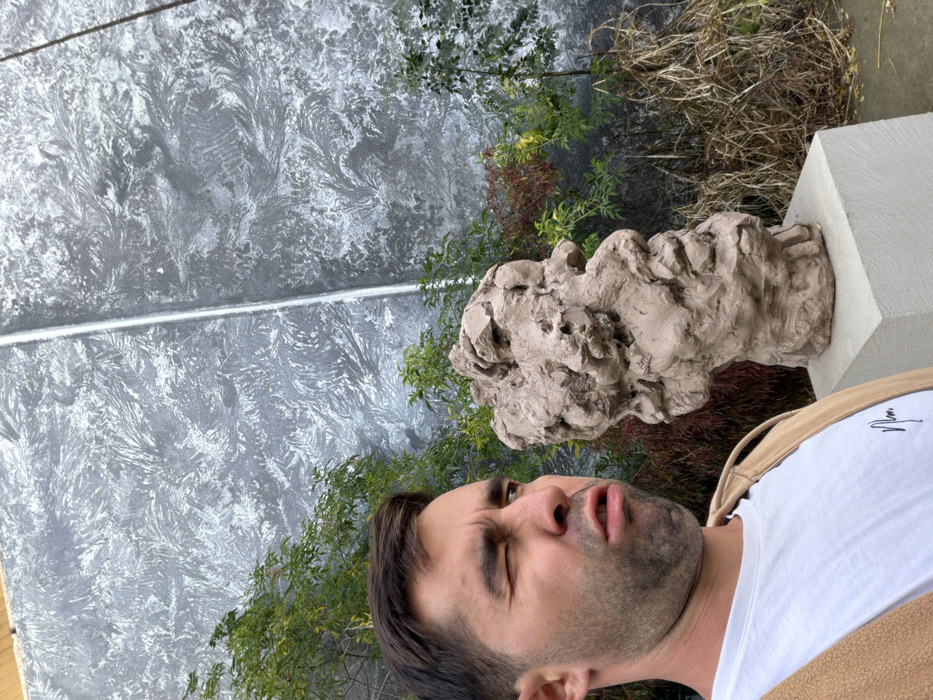 An outdoor scene in Tel-Aviv, Israel, depicts a male individual and a sculpture positioned against a textured wall and natural foliage. The male subject, shown from the chest up on the left, has dark hair and stubble, wearing a white t-shirt with a dark logo and a light brown vest. He exhibits a deliberate facial expression: his left eye is winked shut, his right eye is open, and his mouth is slightly open with pursed lips, indicating a comedic or exaggerated reaction, possibly directed towards the sculpture. To the right of the man, an abstract, light brown sculpture resembling a highly textured and distorted human head or bust rests on a light gray rectangular pedestal. The background features a tall, dark gray wall with a prominent, intricate light gray and white swirling or crystalline pattern, divided by a vertical lighter seam. Various green and dried reddish-brown plants and grasses are visible in front of the wall, behind and around the sculpture and the man.