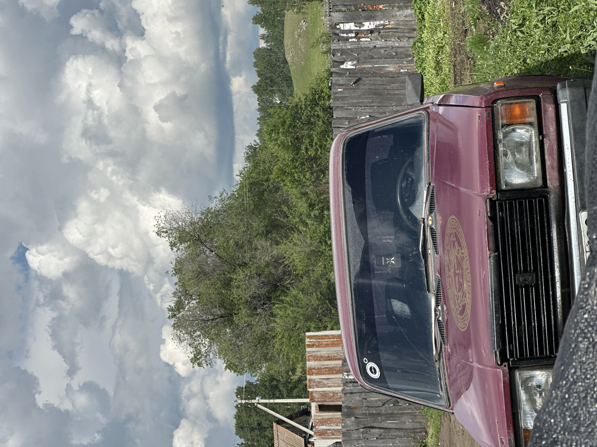 A maroon car is parked on a dirt road in front of a weathered wooden fence. The car's hood is adorned with a circular, golden emblem. The car's windshield has two stickers, one circular with a silhouette and a degree symbol, and another rectangular sticker with black lettering. Beyond the fence, lush green trees and a gently sloping hill are visible under a cloudy sky. In the background, a rustic wooden structure and a utility pole are present. The overall scene depicts a rural or semi-rural setting.