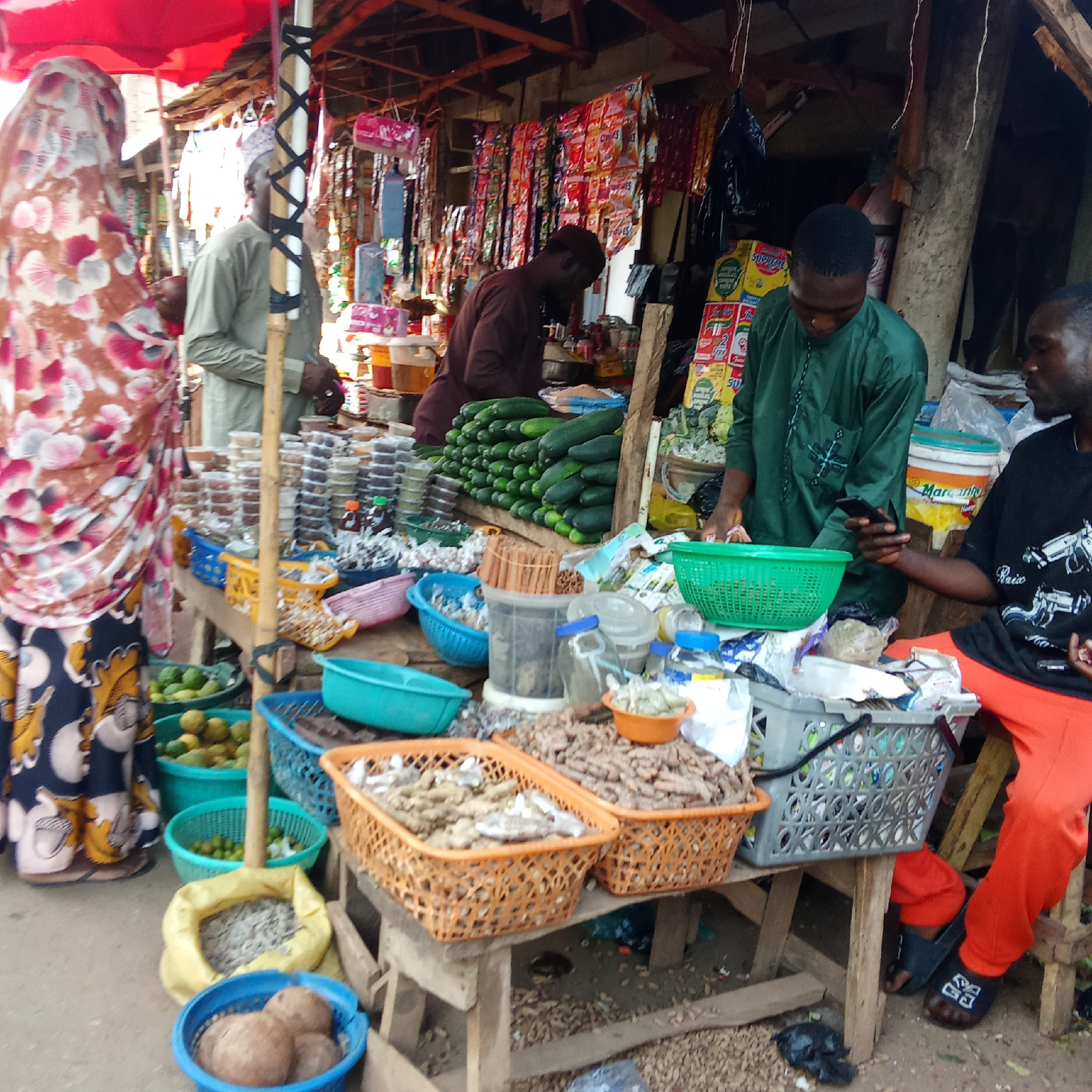 The image captures a bustling market scene in Akwanga, Nigeria. A woman in a patterned headscarf and dress stands near a vendor with rows of goods. Baskets brimming with turmeric, limes, coconuts, and small snacks are arranged on wooden tables. Three men, one in a brown outfit, one in a green outfit, and the third in a black t-shirt and orange pants, are visible behind and to the right of the stand, attending to sales and using a cellphone. The market stall is under a partial awning, with various snacks hanging from the structure.