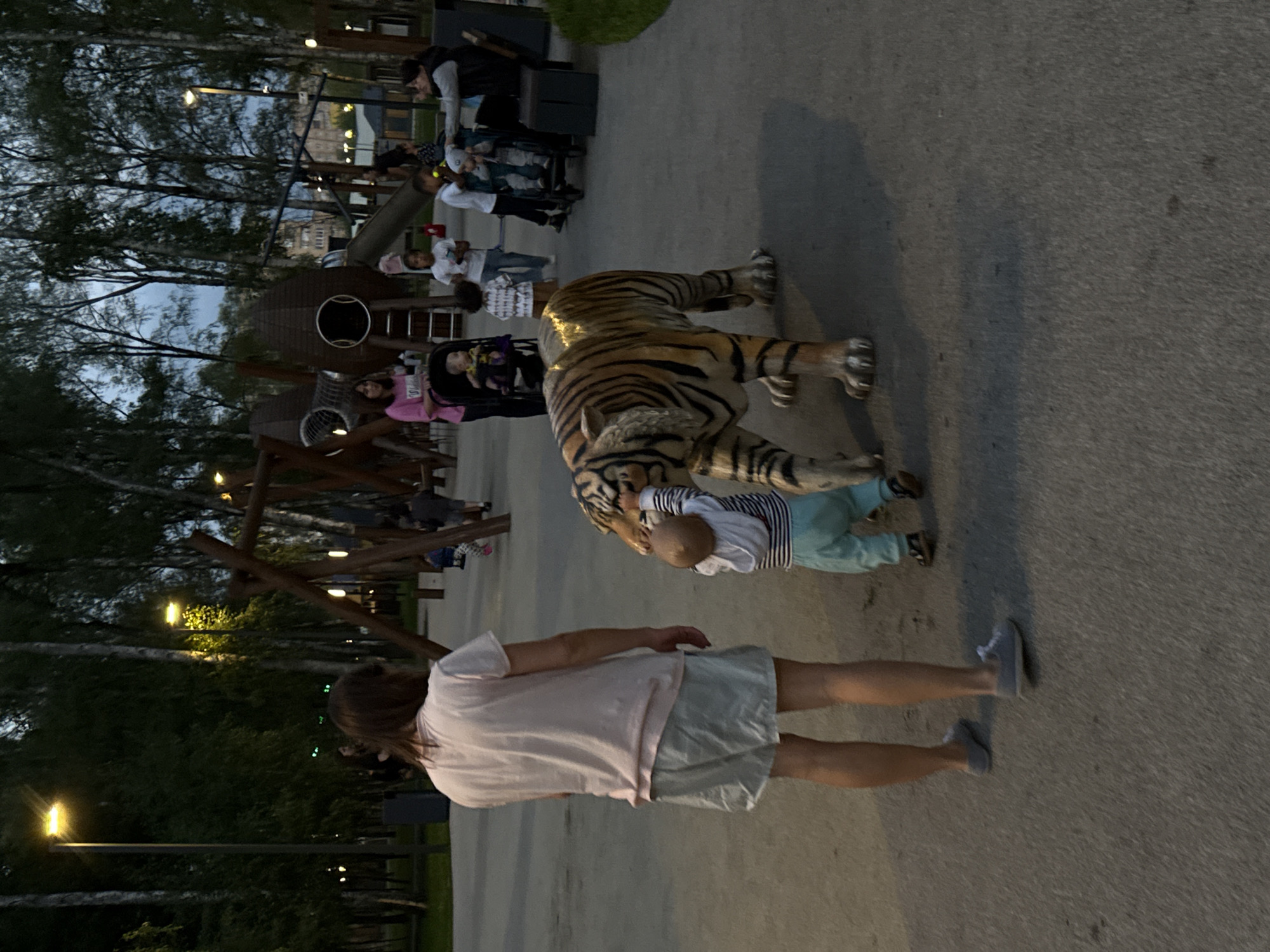 This is a medium-shot, eye-level photograph taken at dusk in the "Gorodskoy Park" in Lobnya, Russia. The scene captures a playground area with children and adults. In the foreground, a large, realistic tiger statue is present. A young child, wearing a striped long-sleeved shirt and teal pants, is interacting with the tiger statue, reaching out to touch its head. To the left of the child and tiger, a woman with long brown hair, wearing a light pink t-shirt and light-colored shorts, stands facing the statue.
In the background, several other individuals are visible. To the right of the tiger statue, another child in a stroller is being attended to by a woman wearing a dark vest over a lighter-colored top. Further back, a group of children and adults are near playground equipment that includes swings and a large, bulbous wooden structure with a slide. The park is illuminated by streetlights, and tall trees with thin trunks, possibly birch trees, line the background. The ground appears to be a paved surface.
