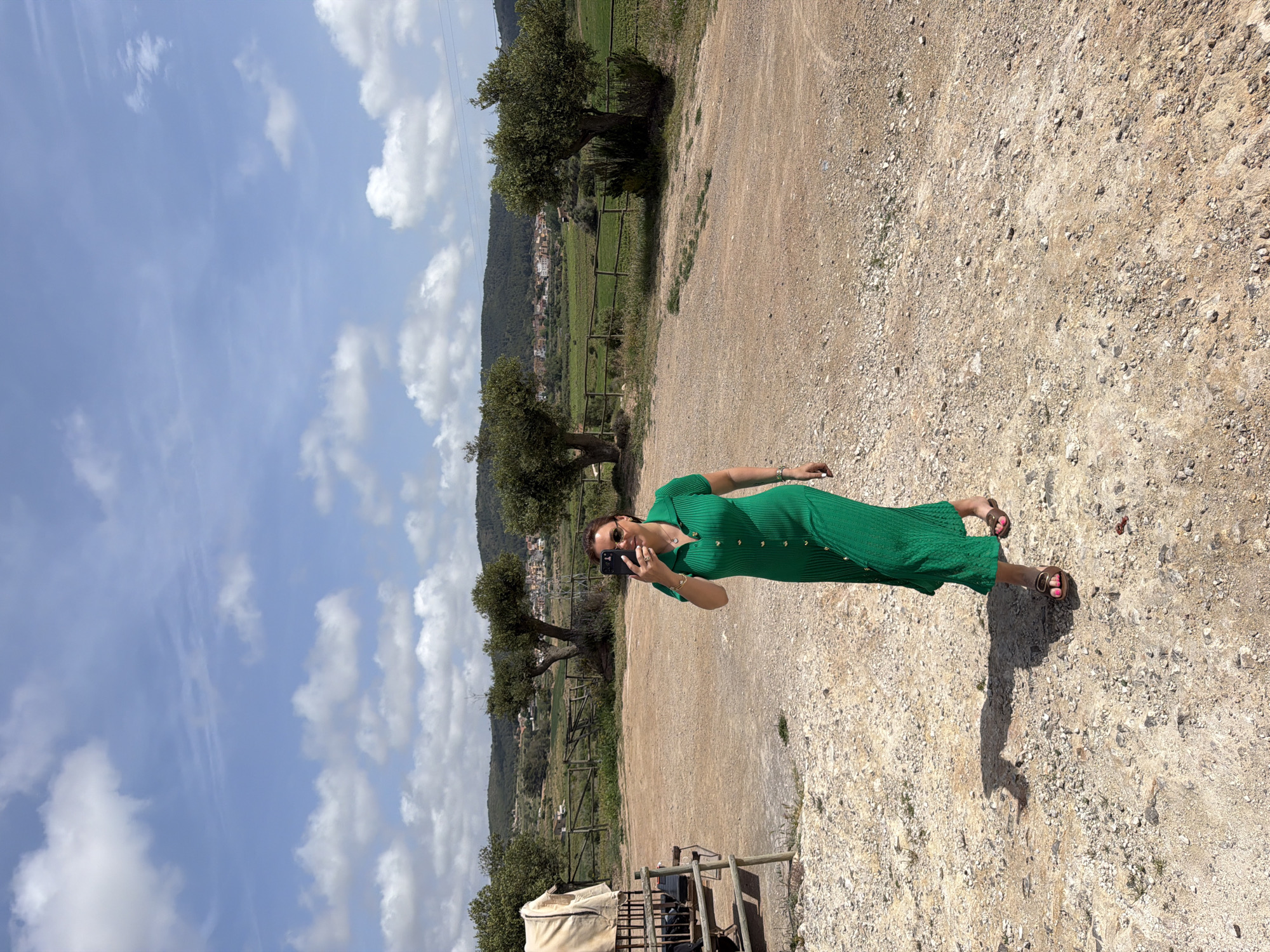 A vertical, eye-level, full shot shows a woman standing on an unpaved, gravel path under a partly cloudy sky in Bonastre, Spain. The bright daylight casts distinct shadows.
The woman, positioned slightly off-center to the right, wears a fitted, short-sleeved, ribbed midi dress in a vibrant emerald green with a collar and gold buttons running down the front. She has dark hair, sunglasses, and is holding a black smartphone up, seemingly taking a selfie or video. She wears light-colored sandals on her feet, and her fingernails and toenails are painted pink.
Behind her, the landscape unfolds into green fields enclosed by wooden fences, scattered with trees, possibly olive trees. In the mid-ground, a small village with light-colored buildings and terracotta roofs is visible, nestled against a backdrop of rolling hills and mountains covered in vegetation. The sky above is a mix of clear blue and scattered white cumulus clouds. The foreground consists entirely of the textured, light brown gravel and dirt path. To the far left, a tent-like structure and some outdoor furniture are partially visible.