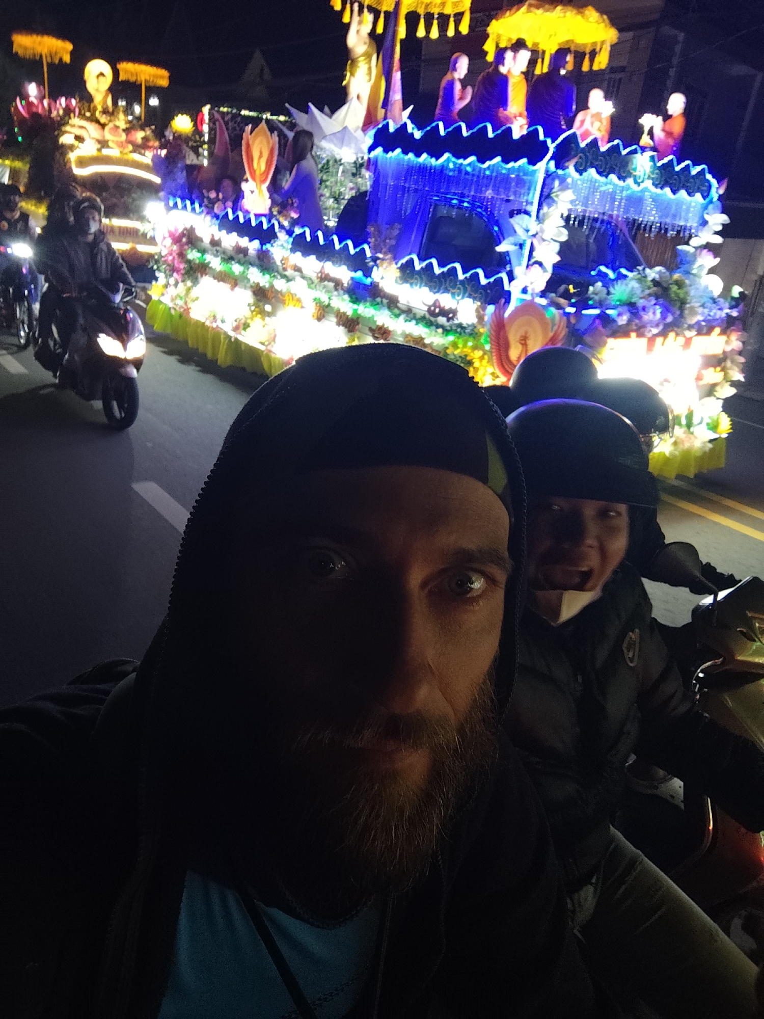 The image is a selfie taken at night in Lang Biang - Đà Lạt, Vietnam, showing two people on a motorcycle. The person taking the selfie is in the foreground, wearing a hooded sweatshirt. A second person is behind them, wearing a helmet and jacket. In the background, illuminated floats with religious figures and decorations are part of a parade. Other motorcycles are also visible. The setting is an urban street at night.