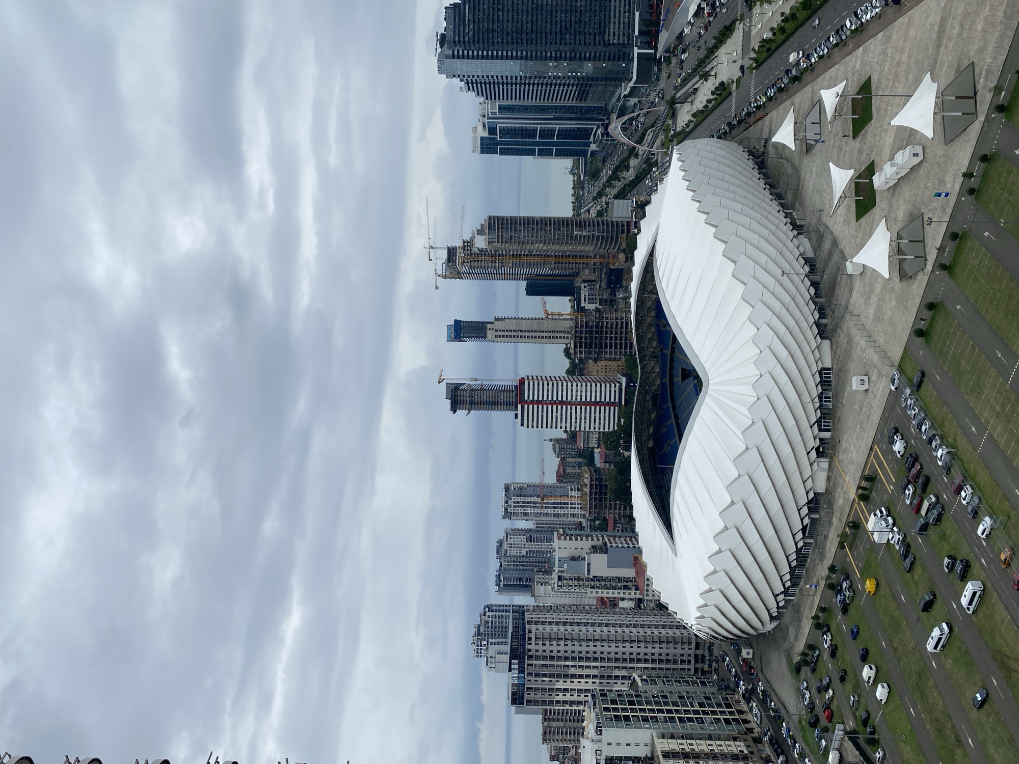 The image captures an aerial view of the Batumi Stadium in Batumi, Georgia, under a cloudy sky. The stadium, with its distinctive white, wavy roof, is the central focus. Surrounding the stadium, a large parking lot is filled with numerous cars, vans, and some white tents or shelters. Beyond the stadium and parking area, a city skyline with numerous high-rise buildings stretches towards the sea. Cranes are visible on several buildings under construction. A road with traffic runs alongside the stadium, leading to a bridge further in the distance. The sea is visible behind the city buildings. The overall scene depicts a modern urban environment with a prominent sports venue.