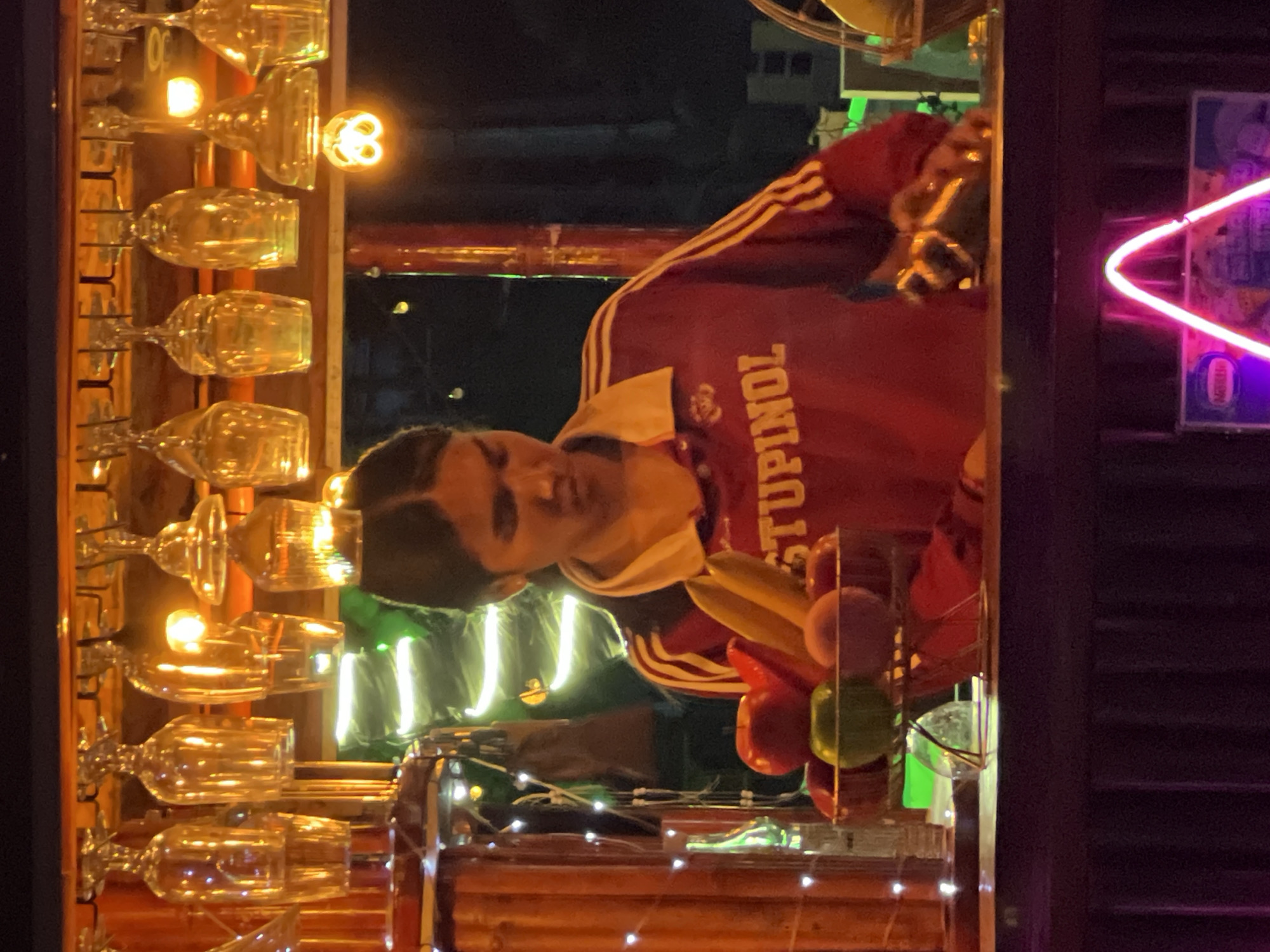A young person with dark hair is standing behind a bar in what appears to be Ko Pha-ngan District, Thailand. They are wearing a red and yellow collared shirt with stripes on the sleeves. The person is looking down, and their hands are resting on the bar. In front of them is a fruit bowl containing apples and a banana. Behind them, various glasses are hanging upside down from a rack. The scene is lit by ambient lighting, including string lights and what appear to be spotlights. A neon sign is visible at the bottom of the frame.
