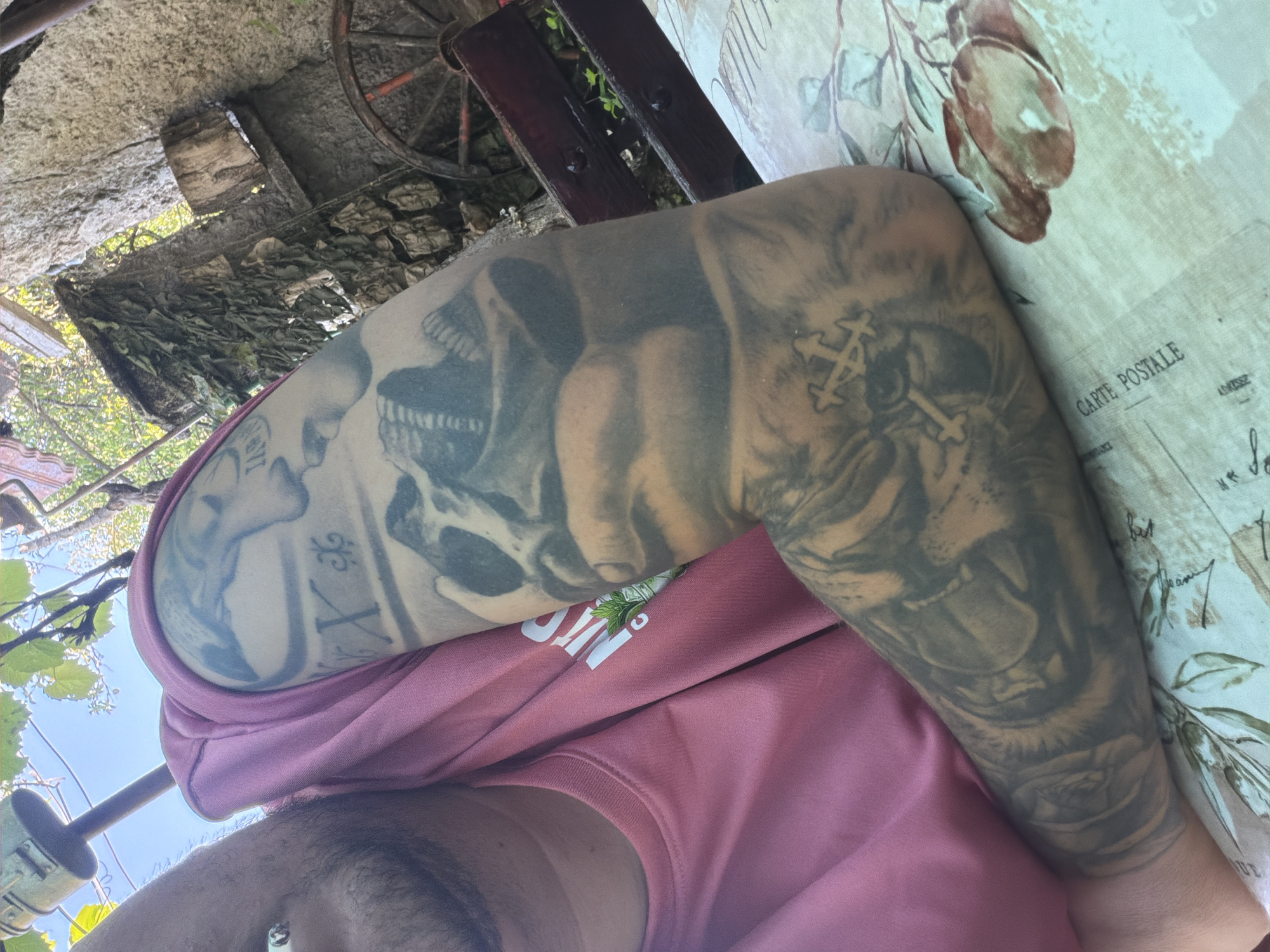 The image shows a close-up view of a person's arm covered in tattoos, resting on a table covered with a tablecloth. The tattoos include a skull, a face, and crosses. The person is wearing a pink t-shirt. The background features a stone wall, foliage, and a rustic wooden object, suggesting an outdoor setting. The scene is set in the city of Kaolinovo, Bulgaria.